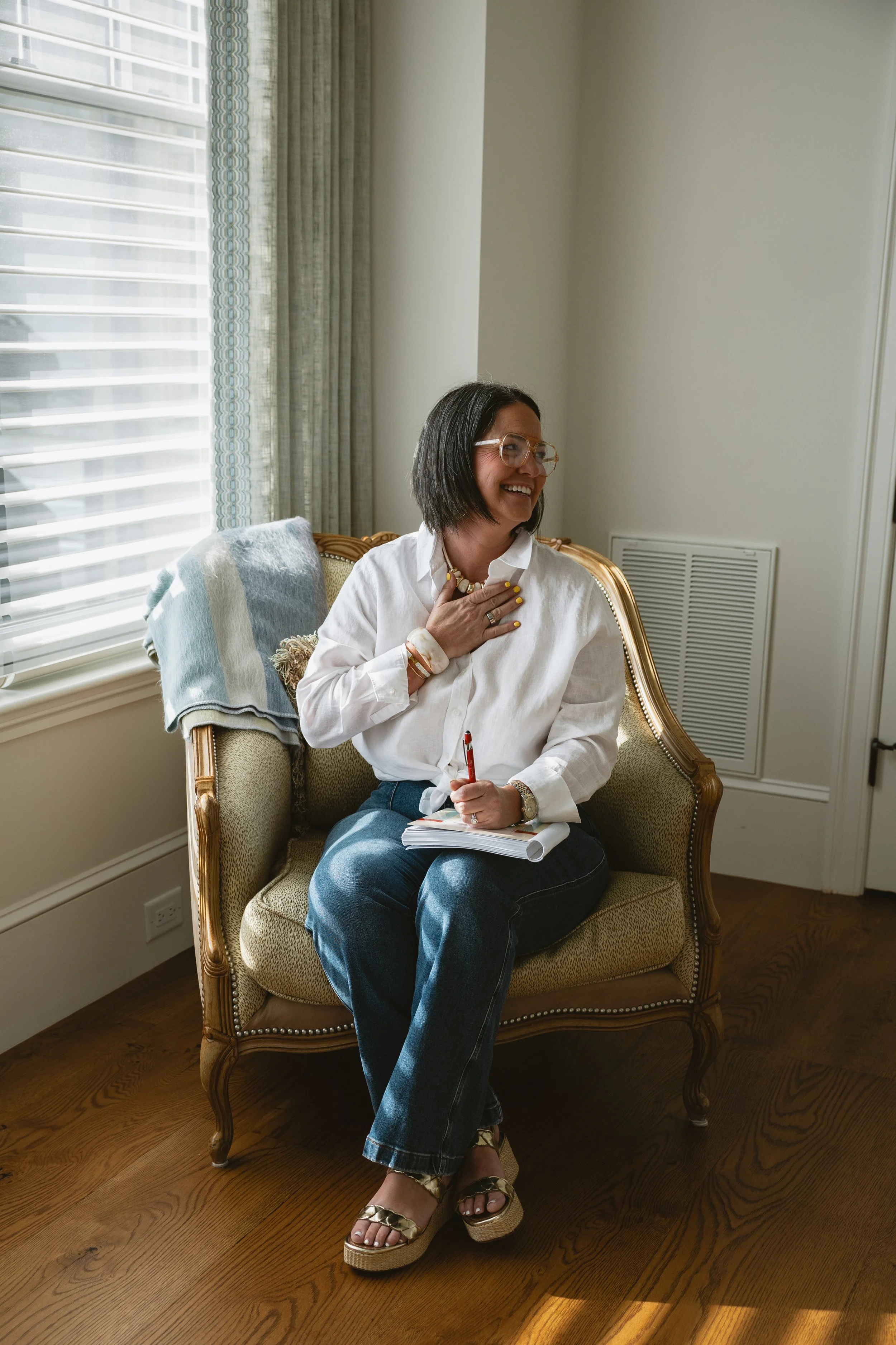 A woman with black hair, glasses, and light skin, sitting on a vintage armchair, smiling, with a hand on her chest holding a necklace, and holding a notebook and pen on her lap, in a well-lit room with large window and wood flooring.