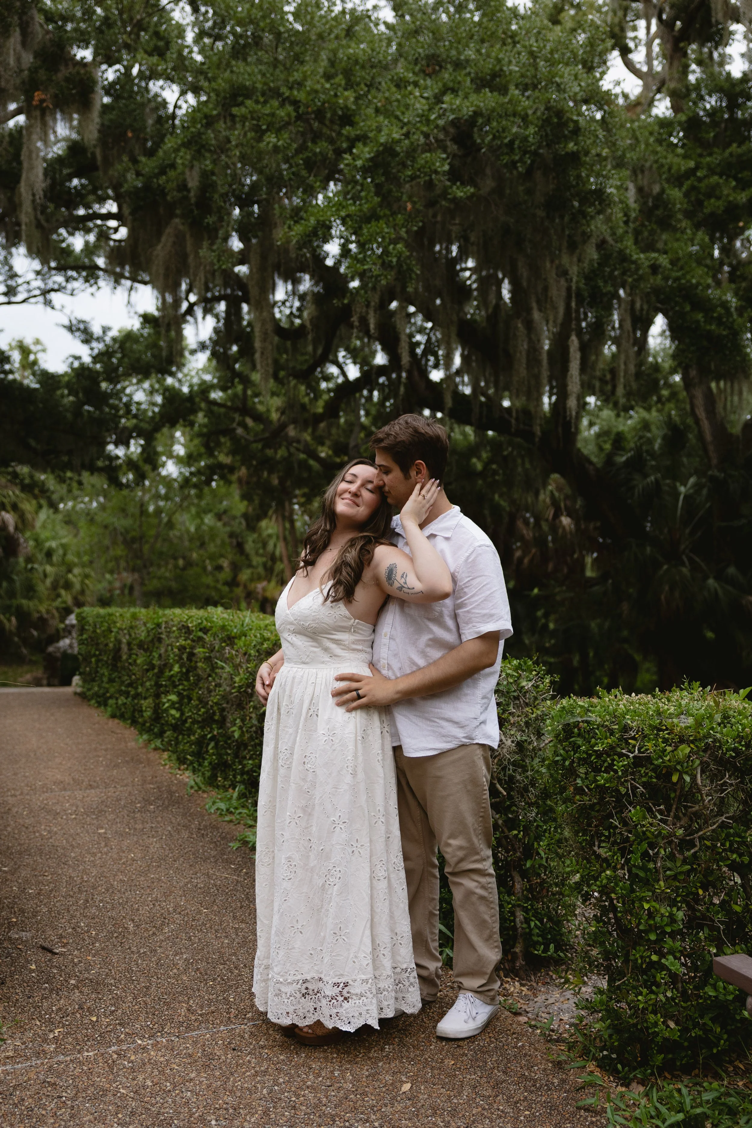 A couple stands close together in a lush, green outdoor garden surrounded by trees and bushes. The woman wears a white, lace, sleeveless dress and the man wears a white shirt and beige pants. They are touching tenderly, with the woman smiling and the