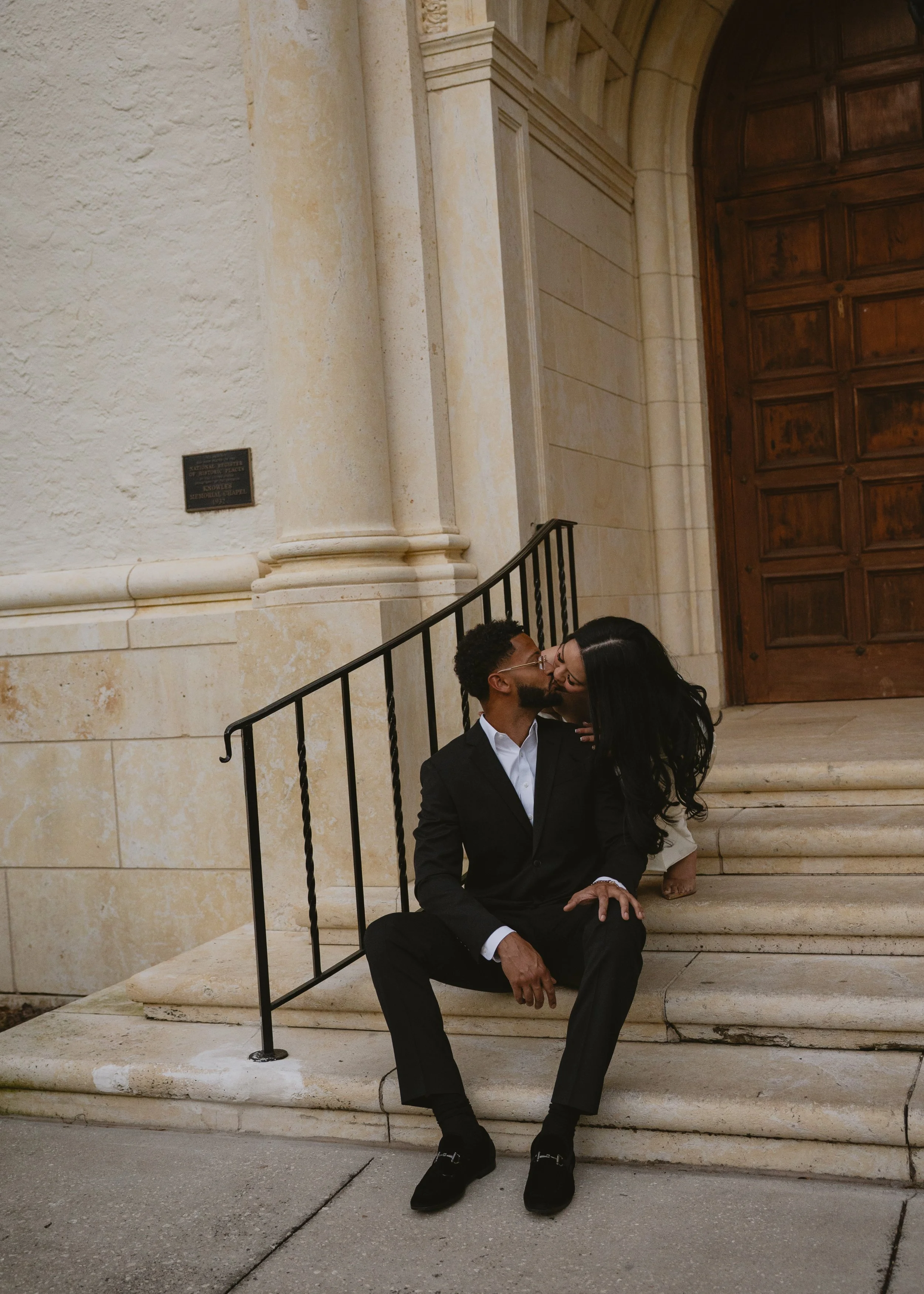 A man and a woman kissing on the steps of a building with large wooden doors, a stone facade, and a small black plaque on the wall.