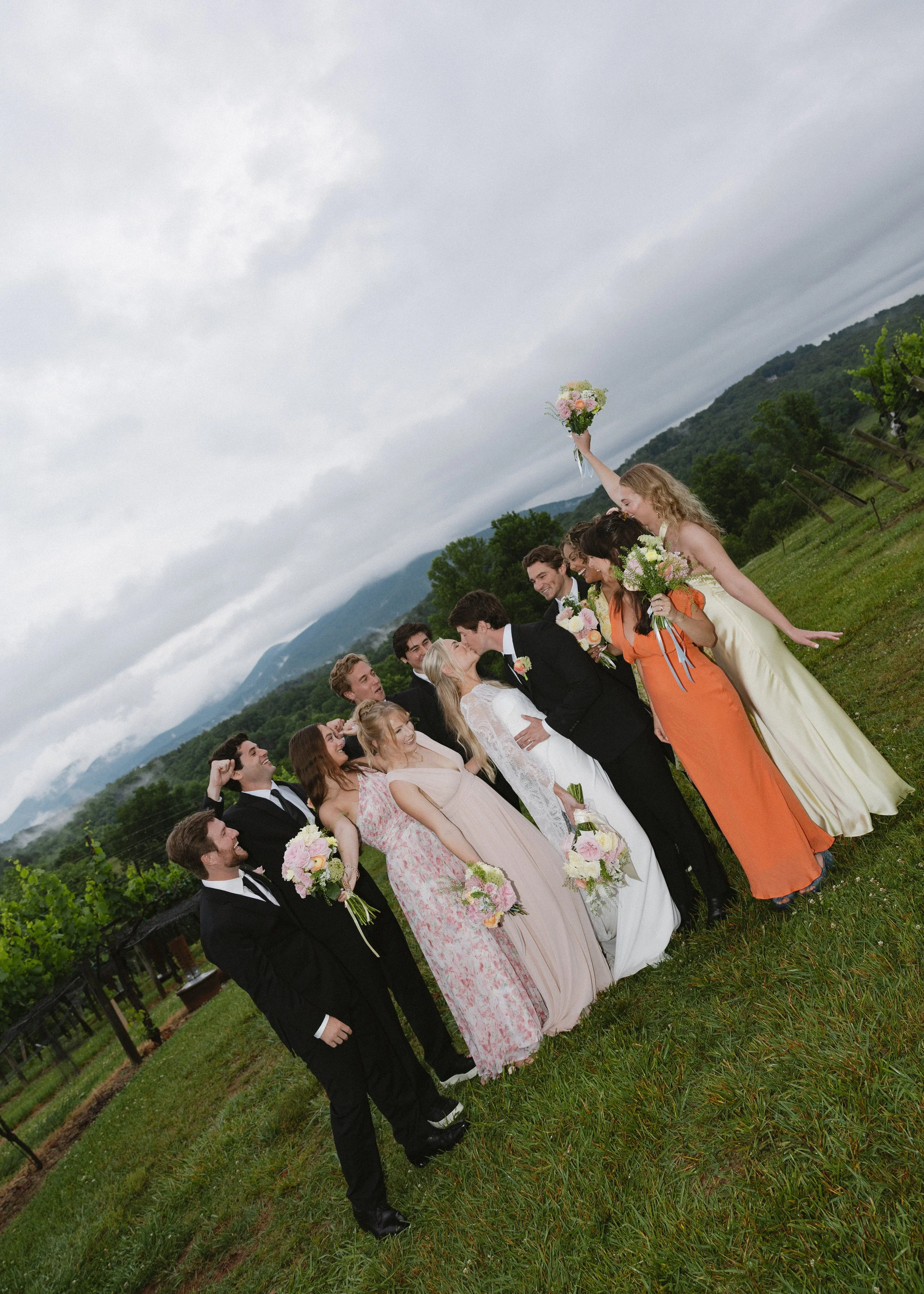 A group of people dressed in formal attire, standing outdoors in a grassy field on a cloudy day, celebrating a wedding with some holding bouquets and others kissing.