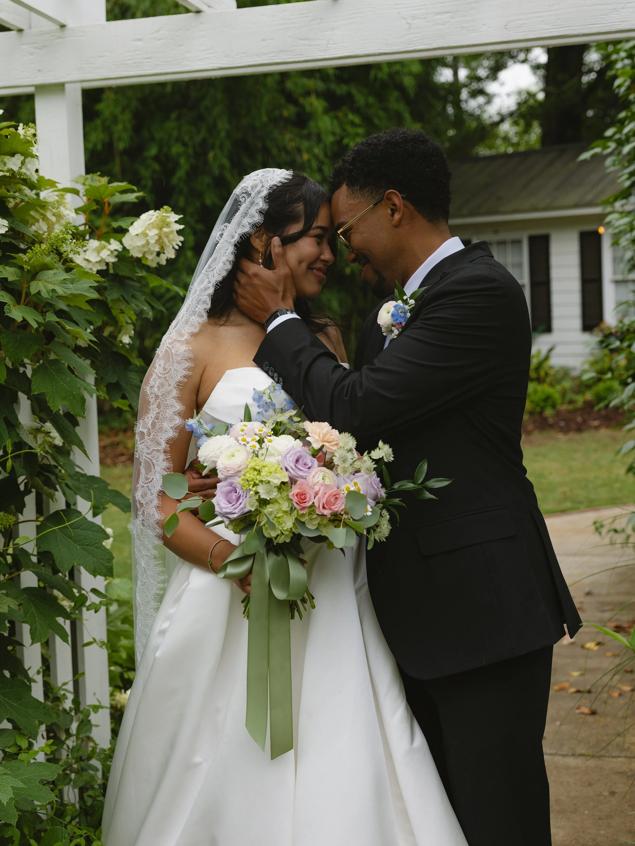 A bride and groom sharing a tender moment during their wedding, with their foreheads touching and eyes closed, outdoors surrounded by greenery and flowers.