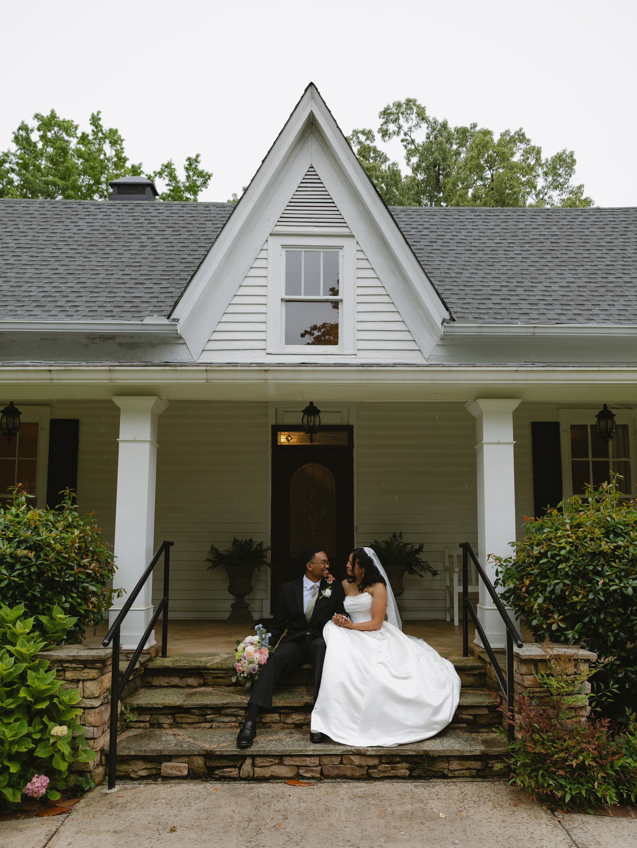 A bride and groom sit on the porch steps of a house, smiling at each other. The bride wears a white wedding gown and veil, holding a bouquet of flowers. The groom wears a black tuxedo with a white shirt and tie. The house has white siding, a gabled r
