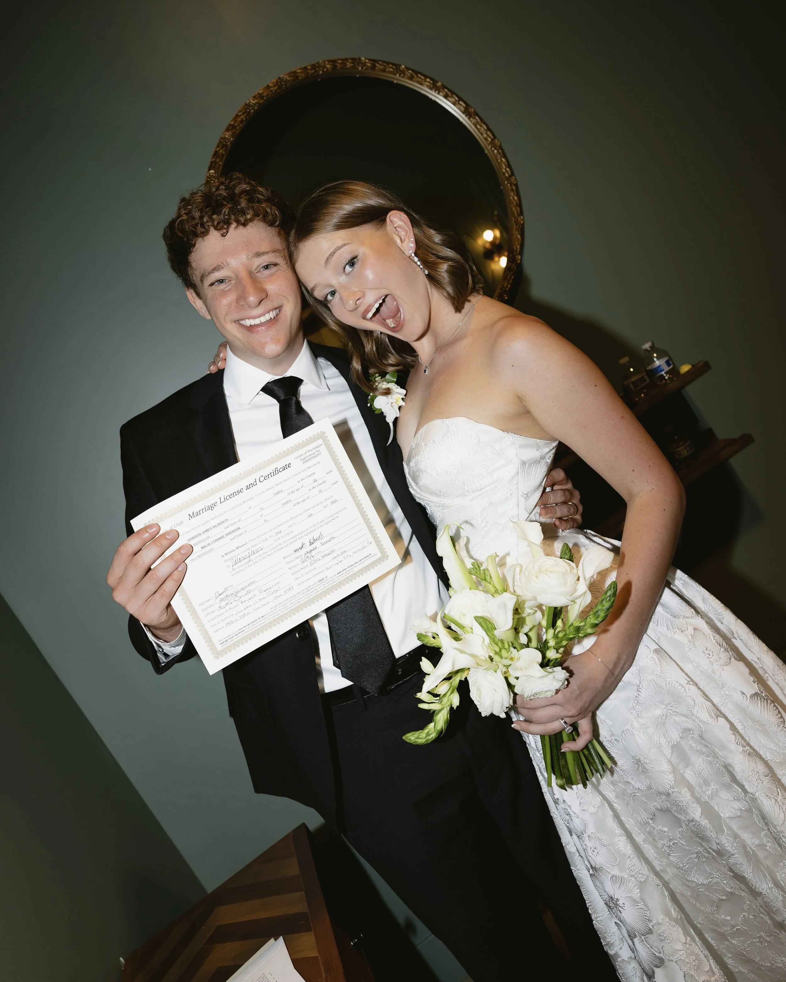A young couple dressed in wedding attire smiling and posing for a photo indoors. The man is wearing a black tuxedo with a white shirt and black tie, holding a marriage license and certificate. The woman is wearing a white strapless wedding gown, hold
