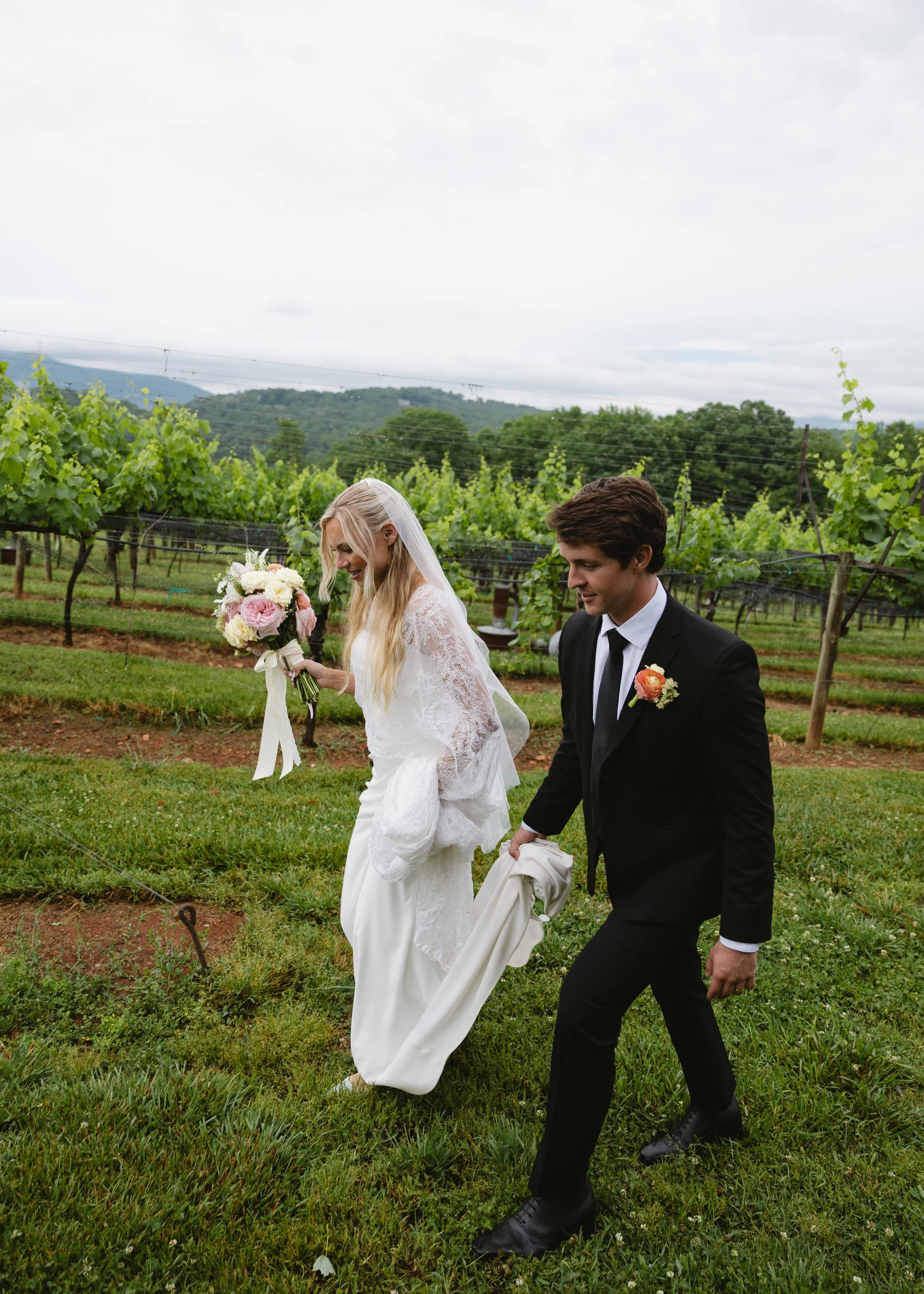 A bride in a white lace wedding dress and veil holding a bouquet of pink and white flowers, walking with a groom in a black suit, white shirt, and tie, during an outdoor wedding ceremony in a vineyard with green vines and hills in the background.