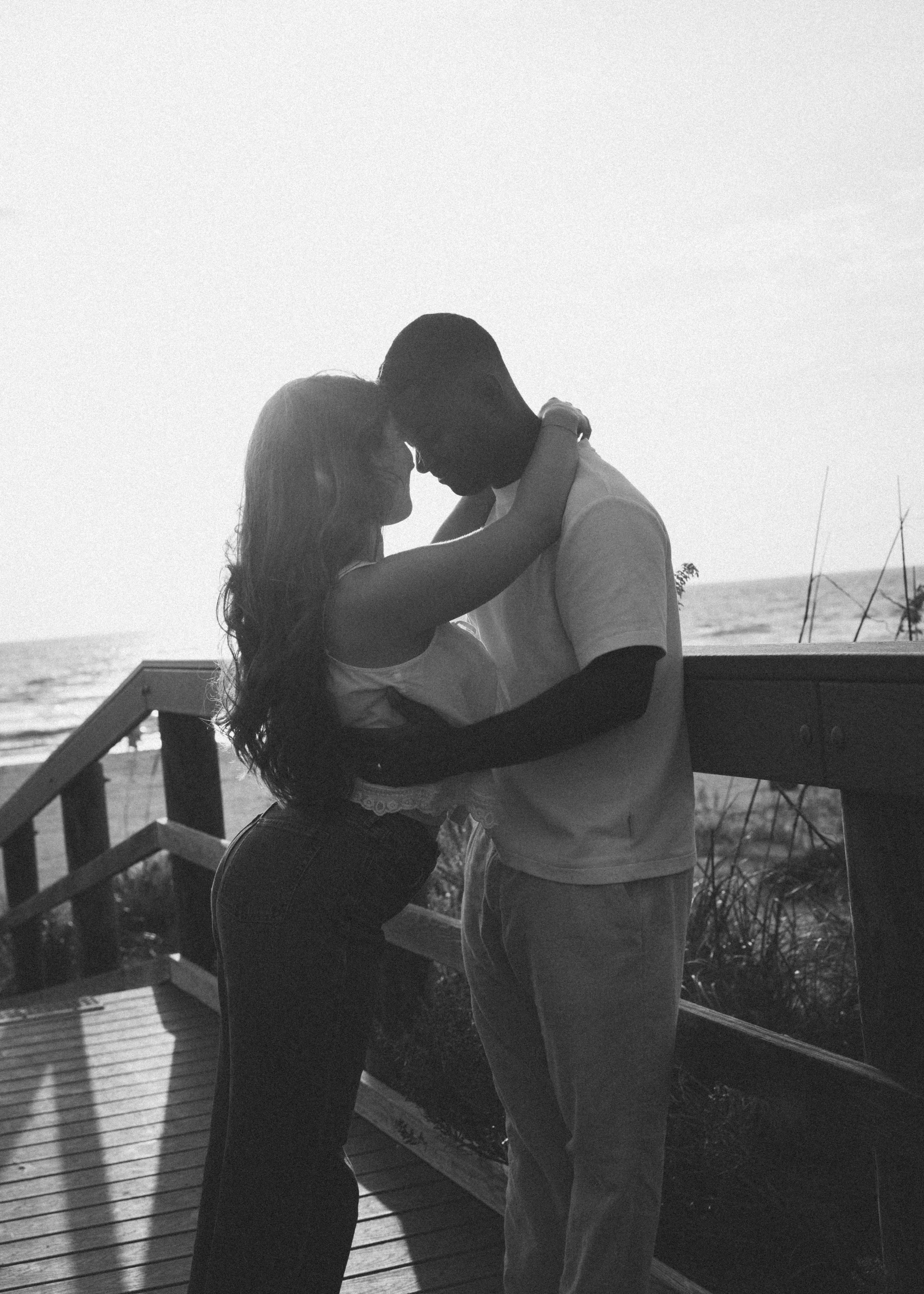 A man and a woman sharing a kiss on a wooden deck by the water at sunset, with the man holding a guitar.