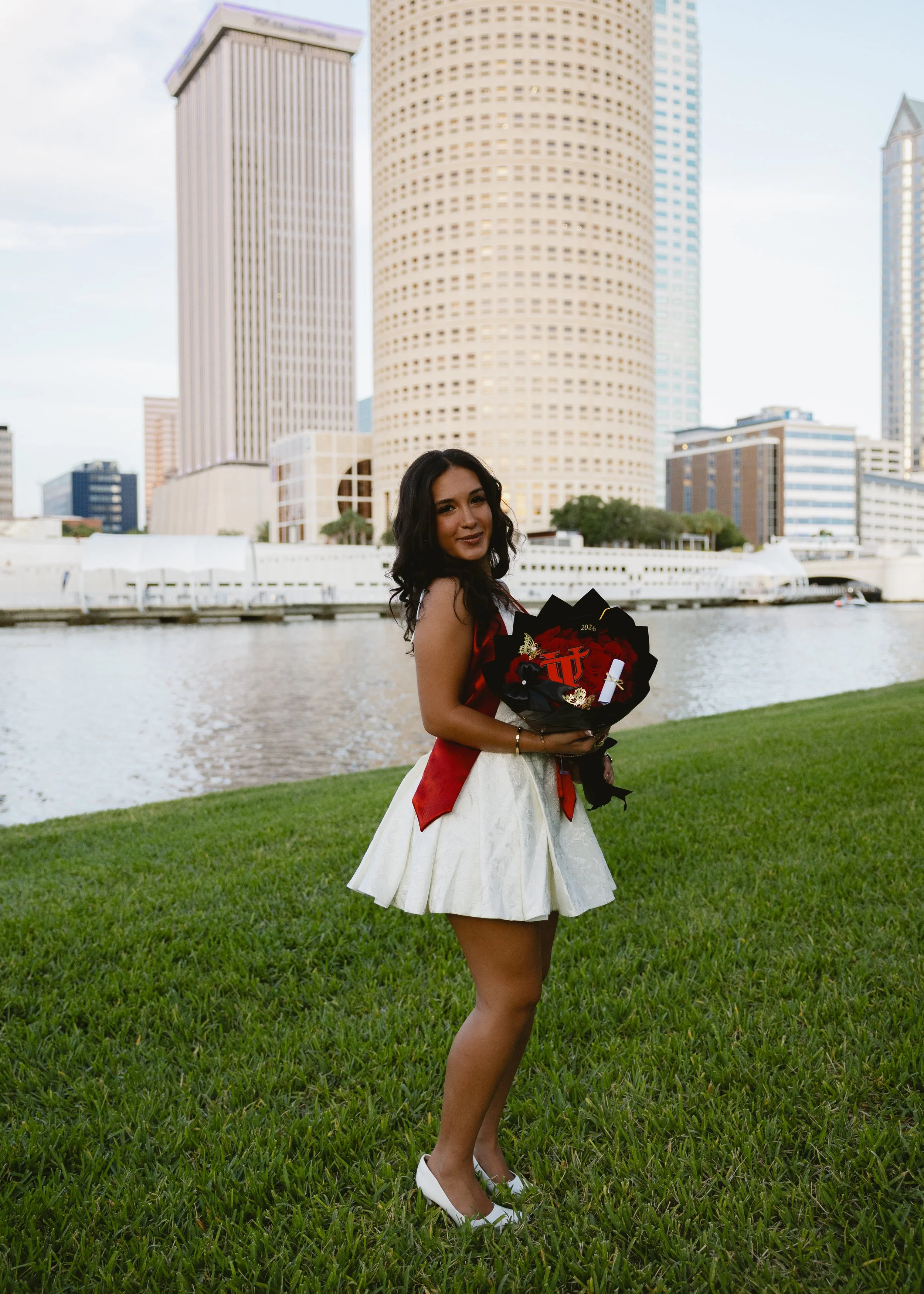 A young woman in a white dress with a red sash is standing on a grassy area near a body of water, holding a bouquet of flowers and a small award. She is wearing white shoes and has dark, wavy hair. The city skyline with tall buildings and skyscrapers