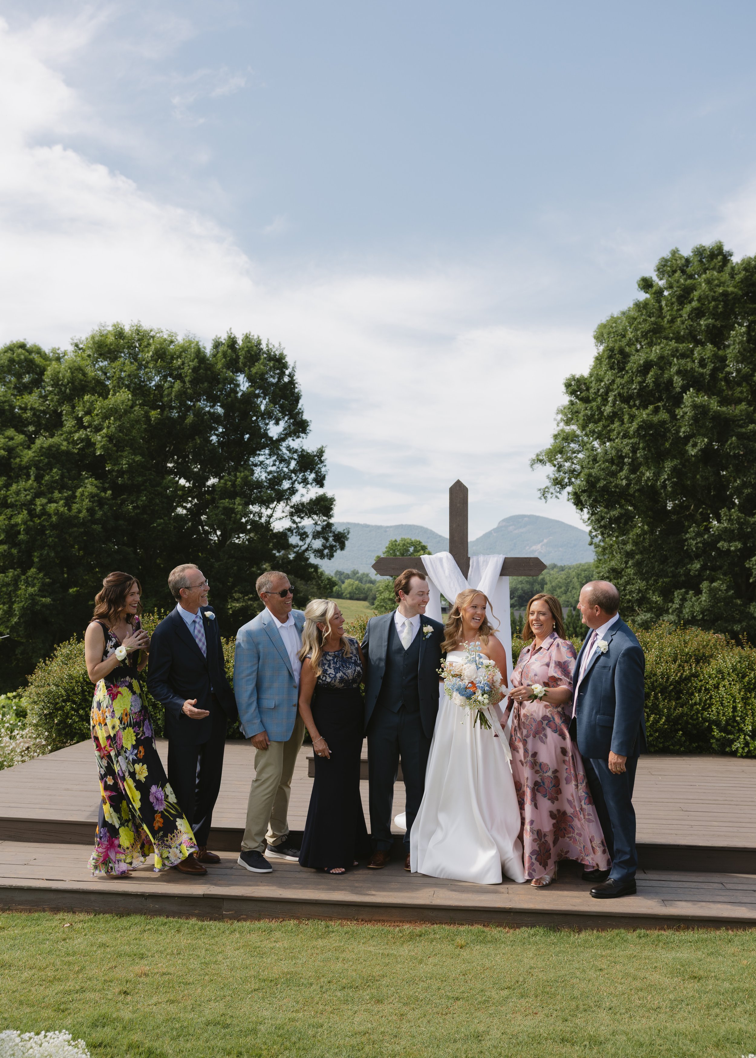 A wedding ceremony outdoors with a group of people standing on a wooden platform. The bride and groom are in the center, smiling and holding hands. The bride wears a white dress and holds a bouquet, while the groom wears a dark suit and tie. Family a