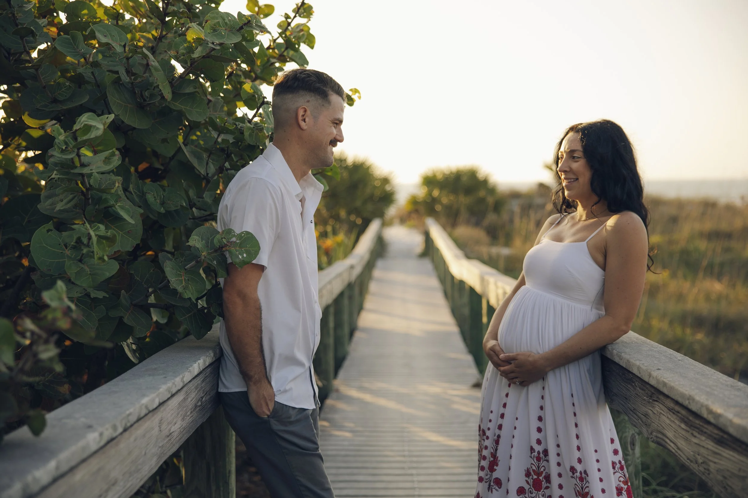 A man and woman are smiling and looking at each other on a wooden bridge during sunset. The woman is pregnant, wearing a white dress with red embroidered patterns, and the man is wearing a white shirt and gray pants. Green foliage and trees are visib