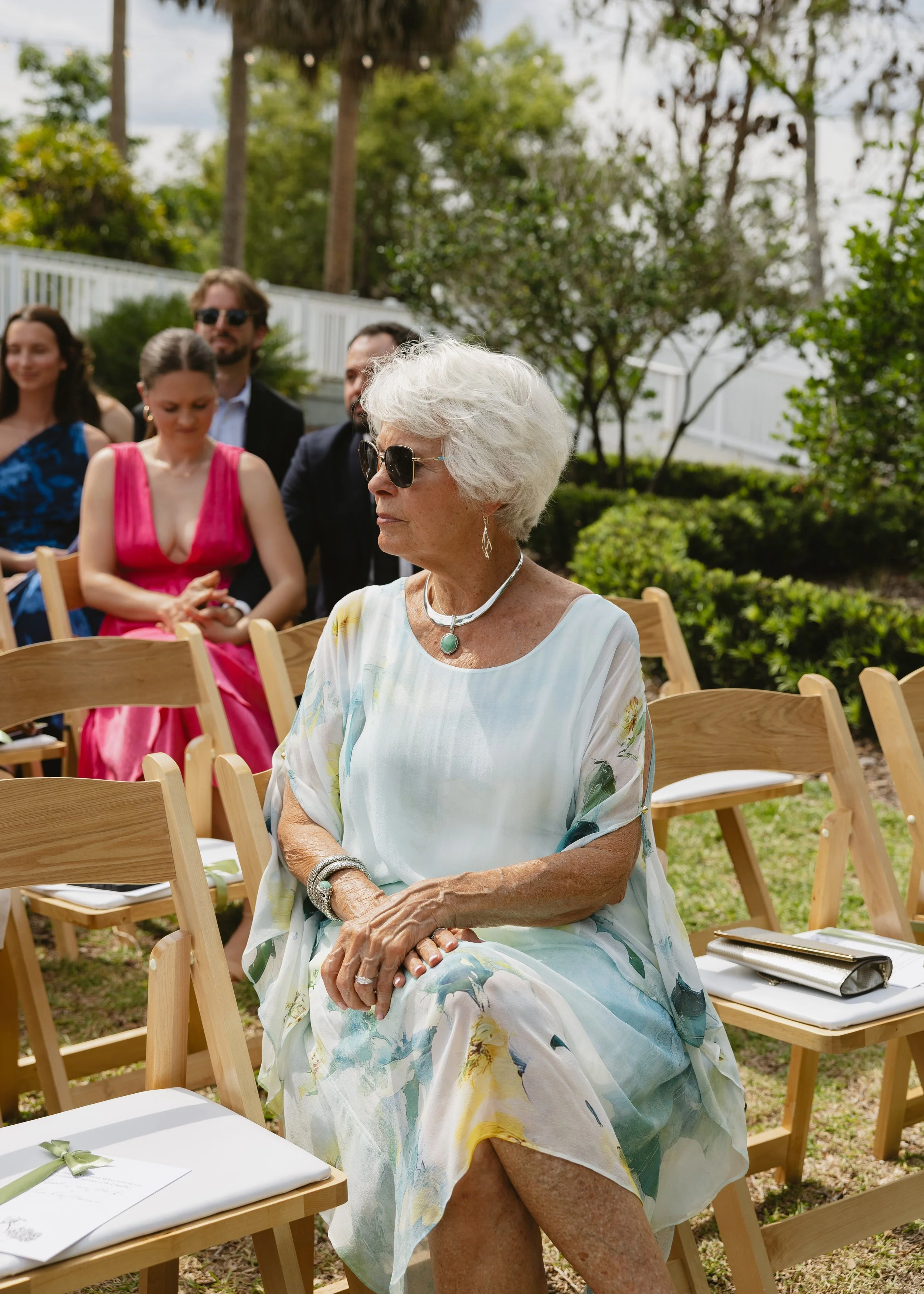 An elderly woman with white hair, wearing sunglasses, a floral dress, and jewelry, sitting outdoors at a daytime event with other people in the background.