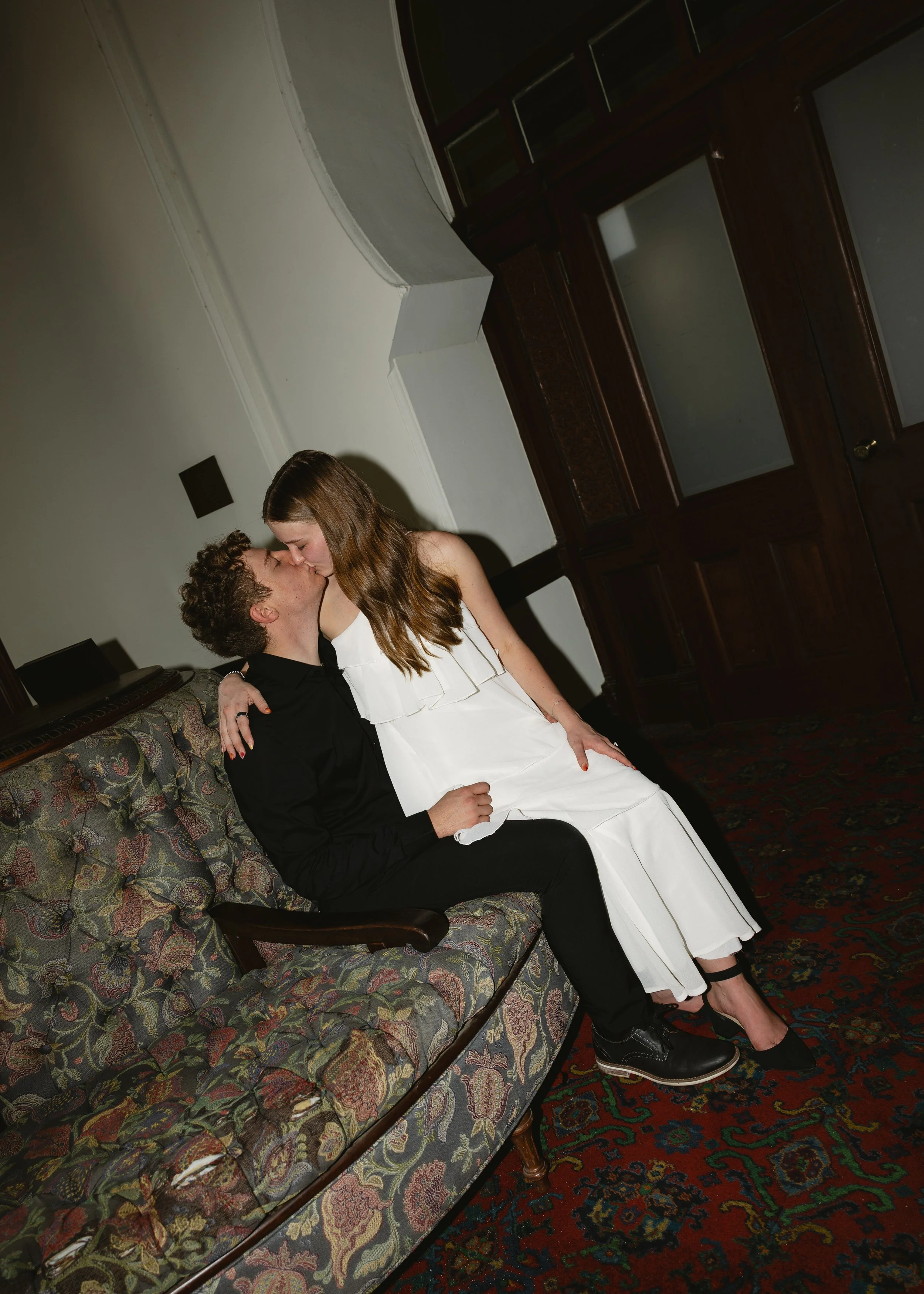 A couple is sitting close together on a vintage floral-patterned couch, sharing a kiss in an indoor setting with dark wooden furniture and a patterned carpet.