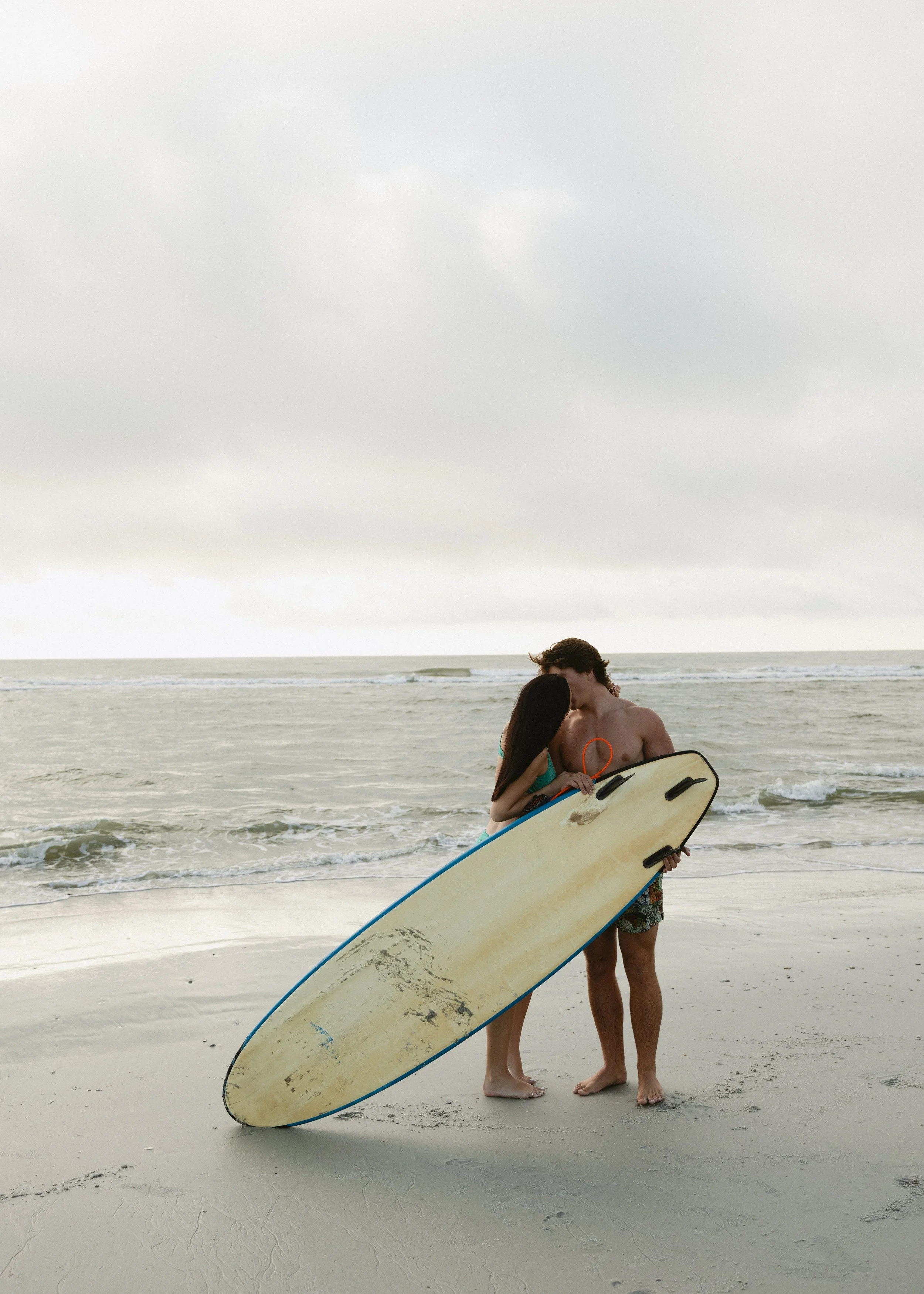 A man and a woman standing close together on a sandy beach, holding a surfboard, with the woman kissing the man's cheek under a cloudy sky.