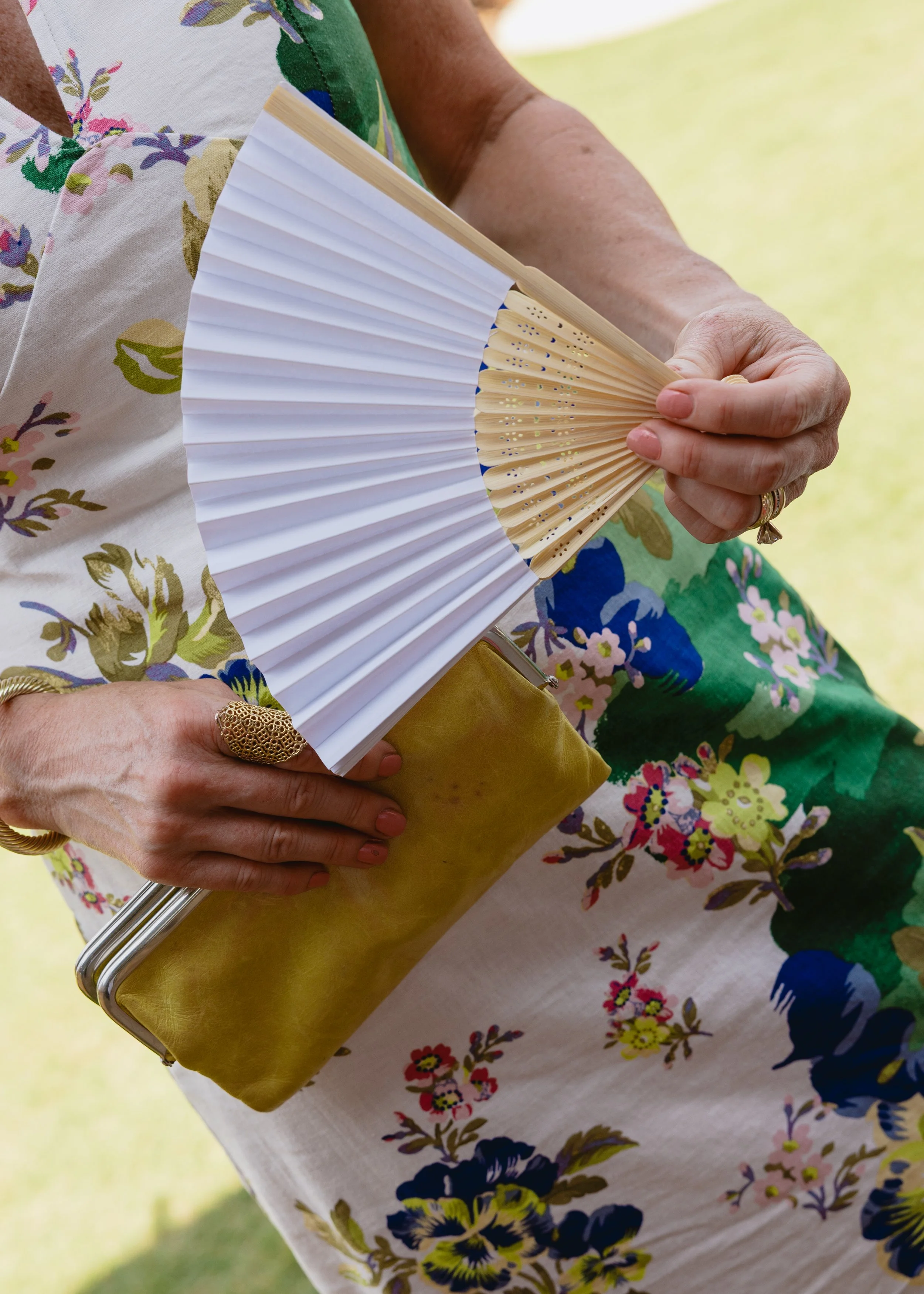 Close-up of a woman holding a white folding hand fan. She is wearing a floral dress with pink, green, blue, and white patterns. Her left hand has a gold ring and a diamond ring. She is holding a gold clutch purse.