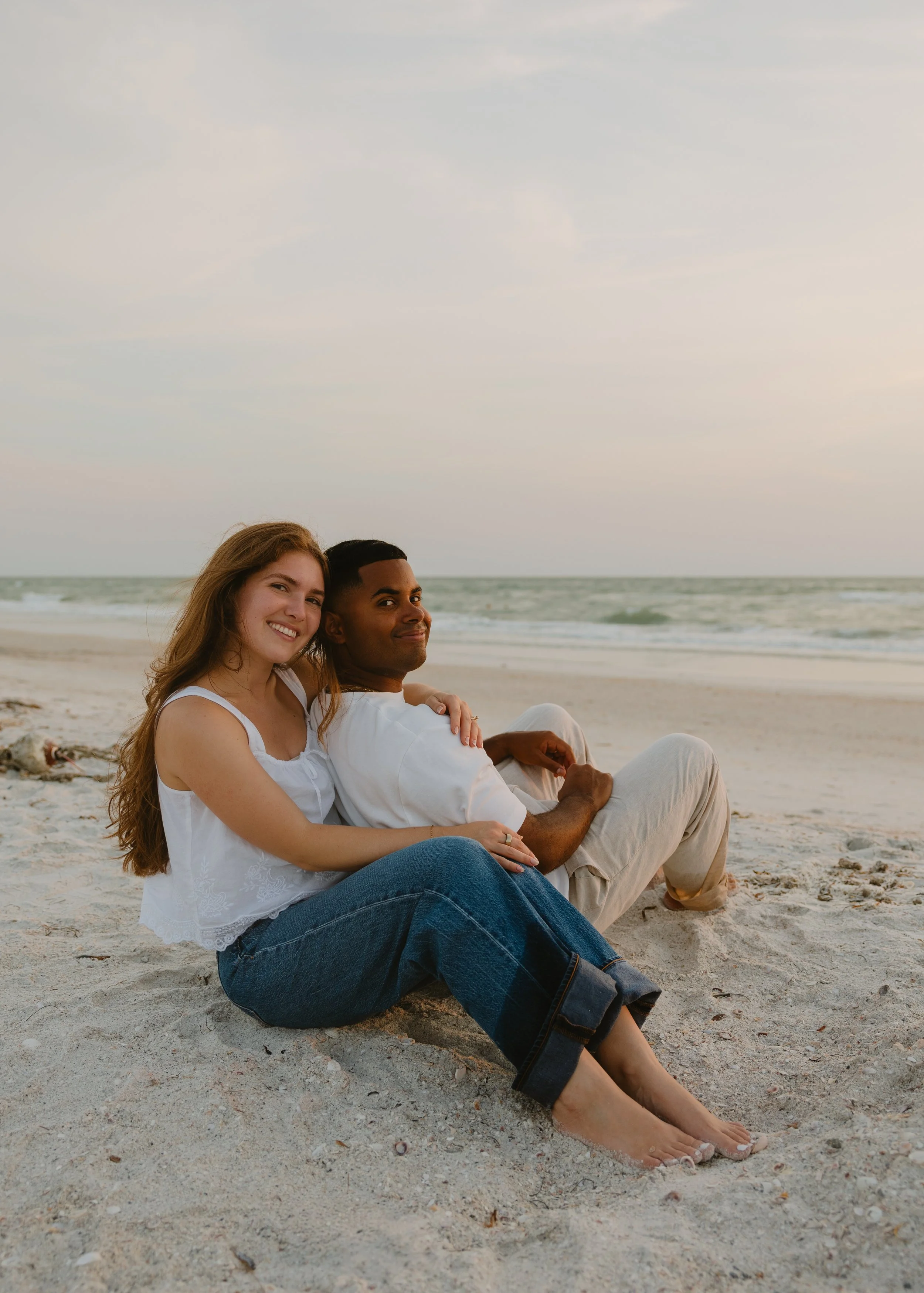 A young couple sitting on the beach with the ocean in the background, smiling and embracing.