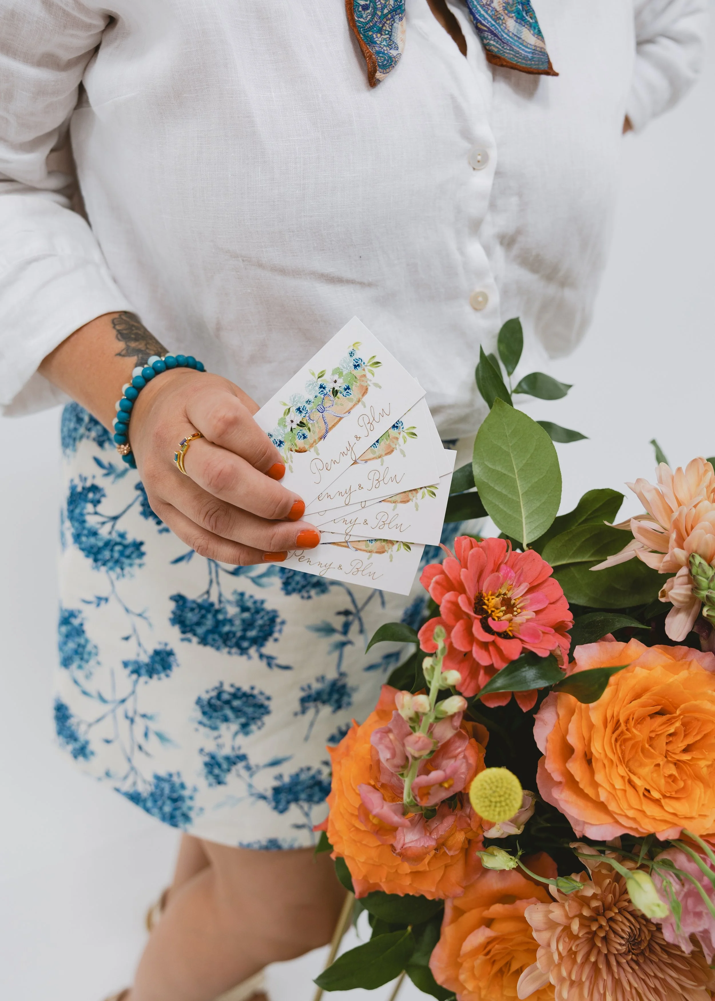 Person holding a bouquet of colorful flowers, wearing a white button-down shirt with a blue patterned skirt, and holding several cards with floral designs and the words "Penny & Blu."