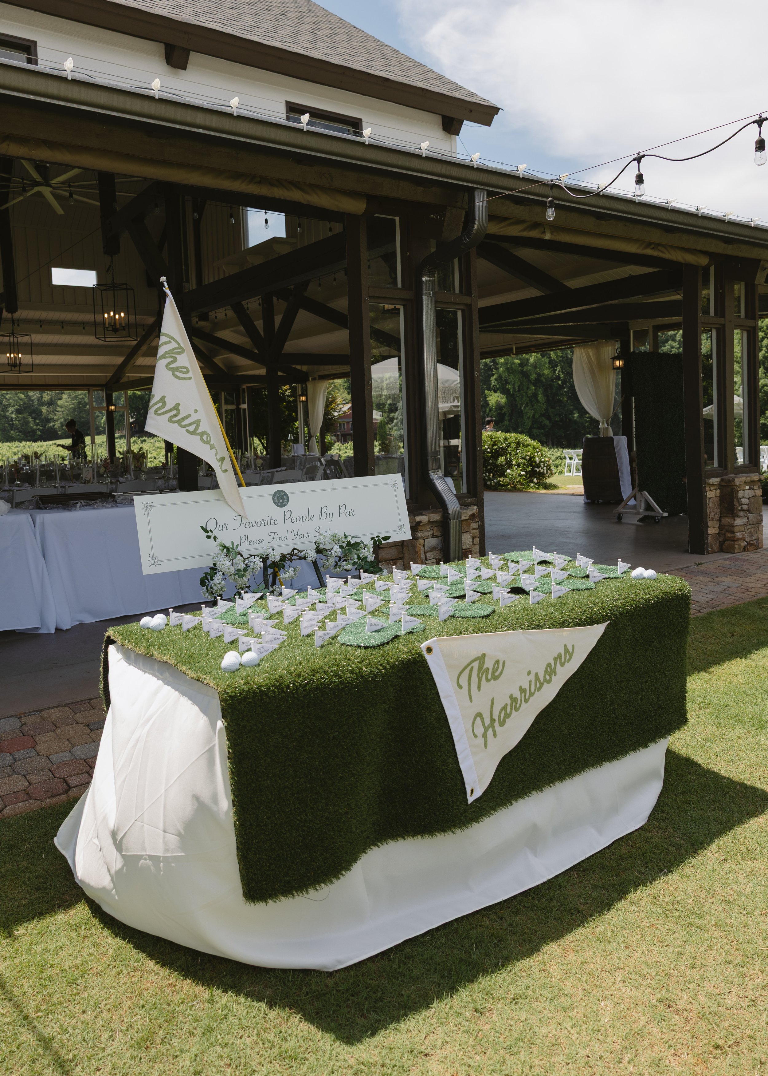 A wedding reception table decorated with a white cloth and a green grass-like centerpiece with small flags that say "The Harrisons." The background shows an outdoor area with a building and string lights overhead.