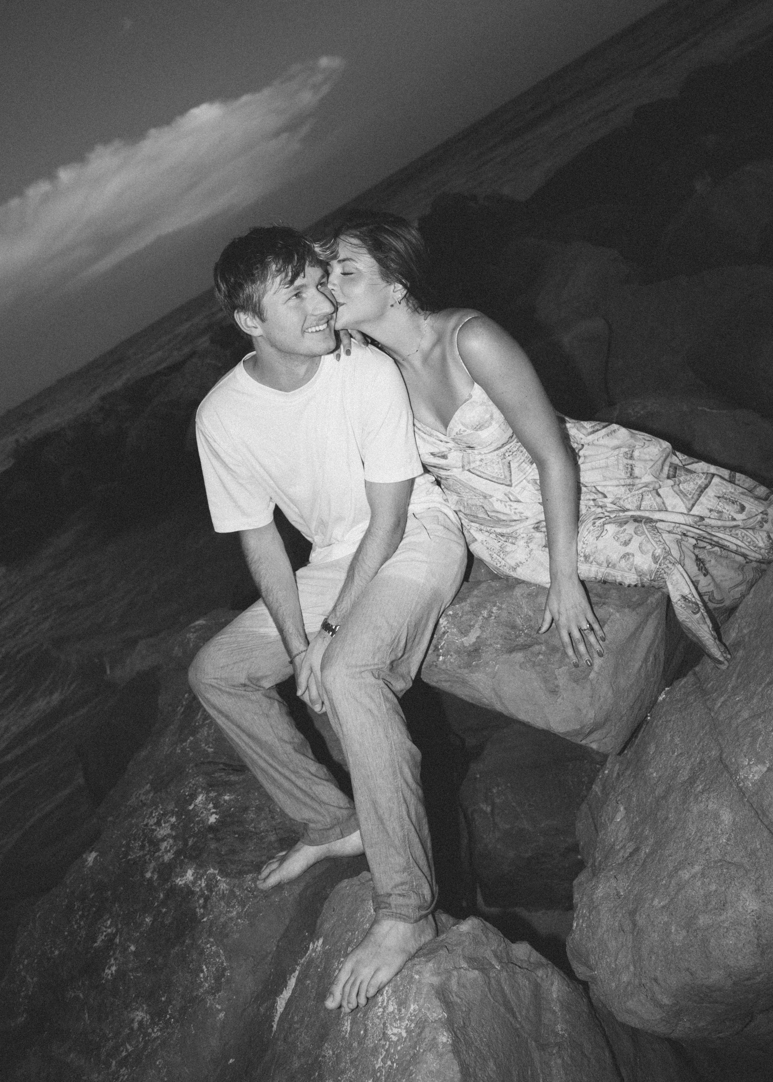 A black and white photo of a young couple sitting on rocks at a beach, with the woman kissing the man's cheek. The man is smiling, wearing a white t-shirt and light-colored pants, while the woman in a patterned dress with a sleeveless top has her han