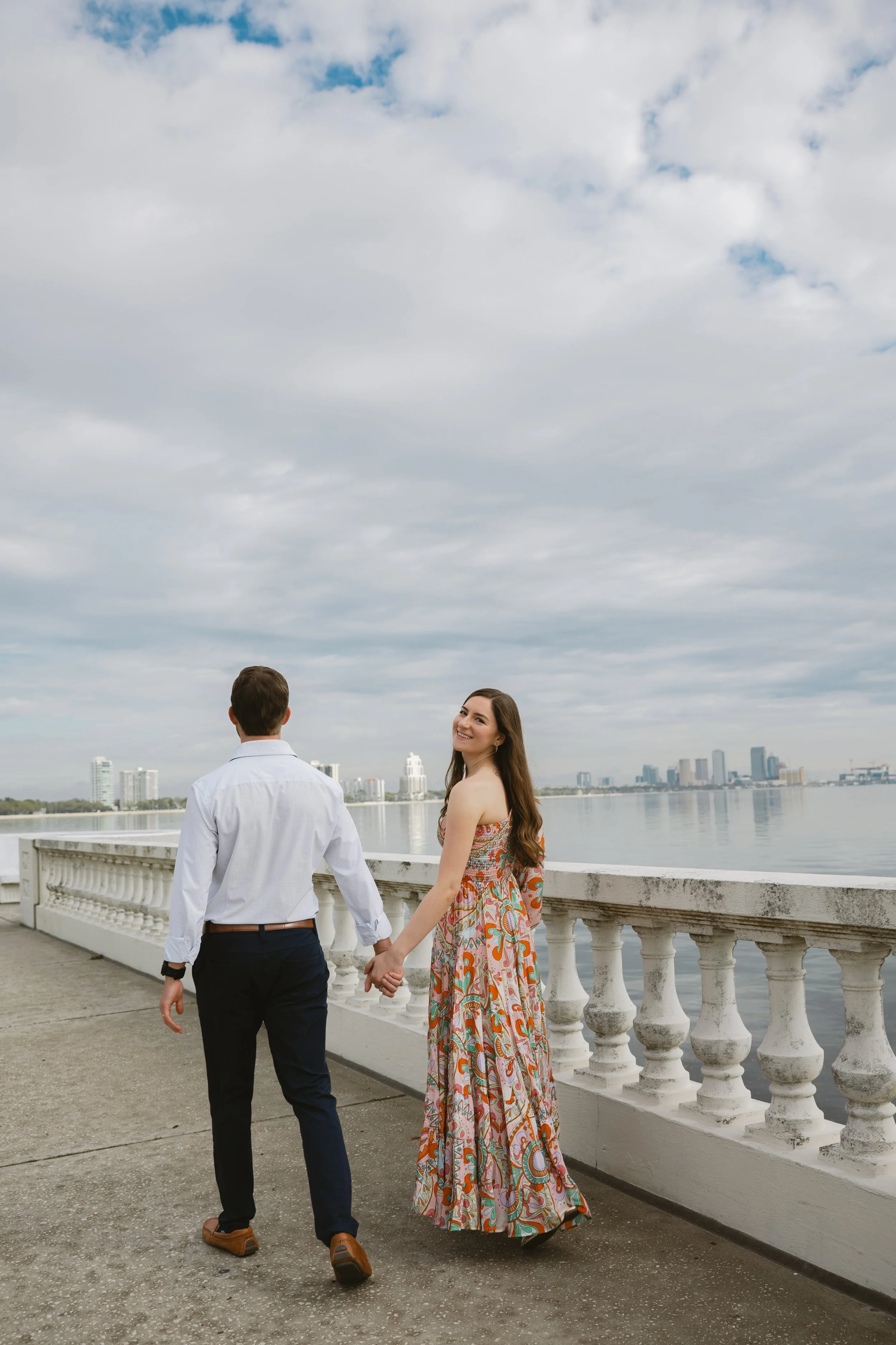 A couple holding hands and walking along a riverside promenade, with a city skyline in the background under cloudy skies.