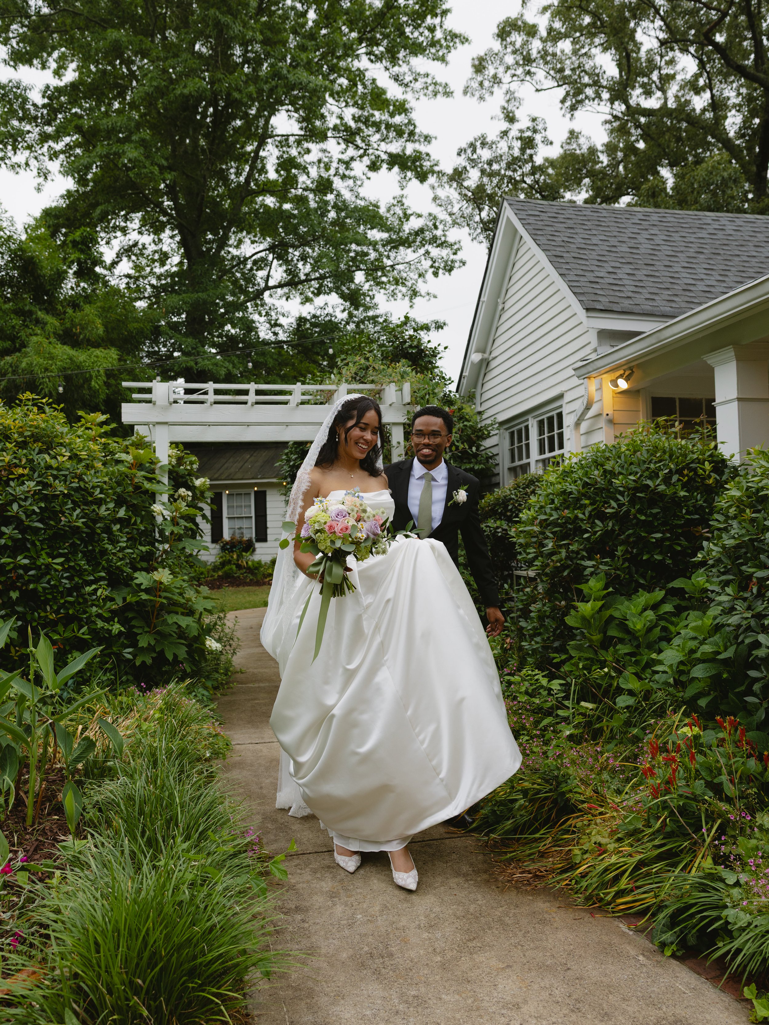 A bride and groom walking outside on a garden path, smiling and holding hands. The bride is wearing a white wedding dress and veil, carrying a bouquet of flowers. The groom is in a dark suit and tie. There are lush green bushes and trees, with a whit