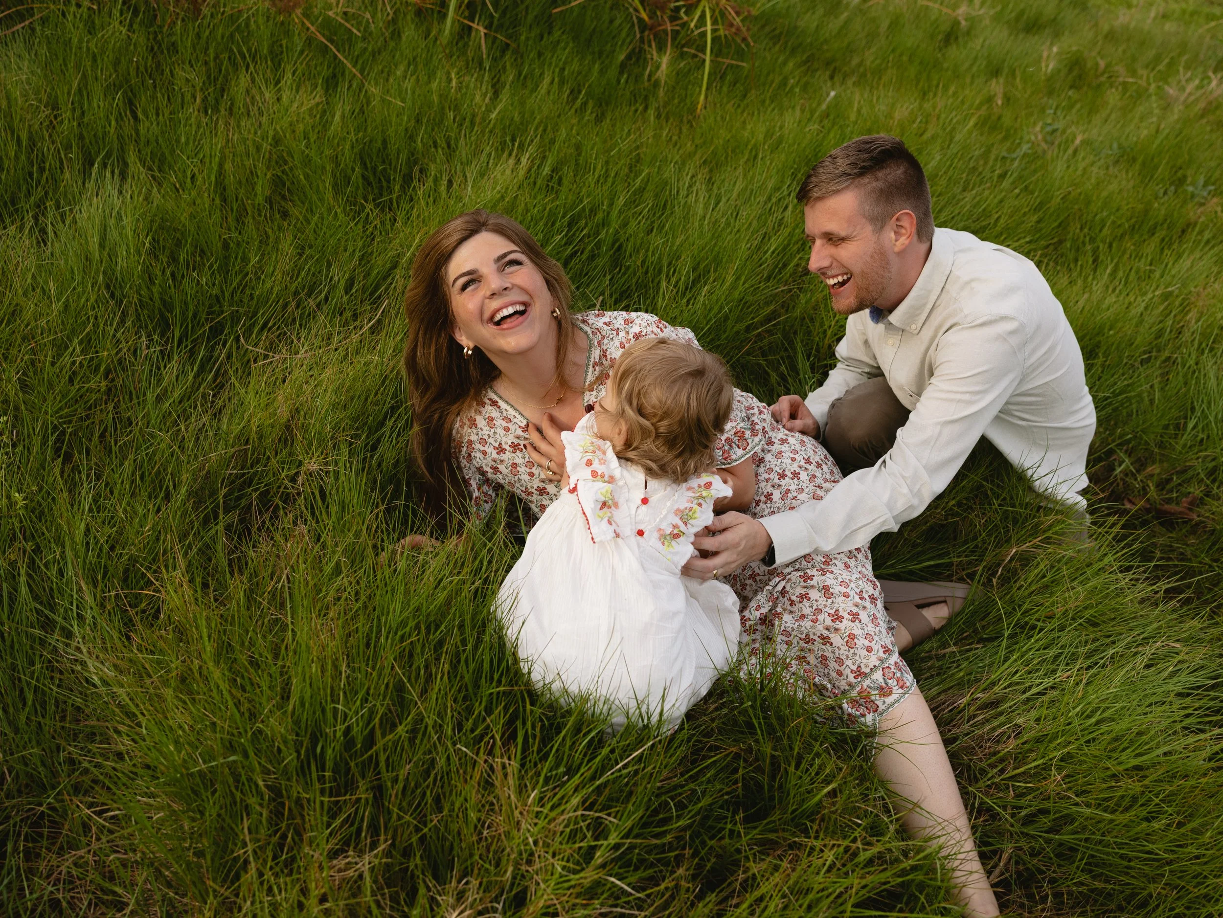 A family of three, including a woman, a man, and a young girl, sitting and laughing in a field of tall green grass.