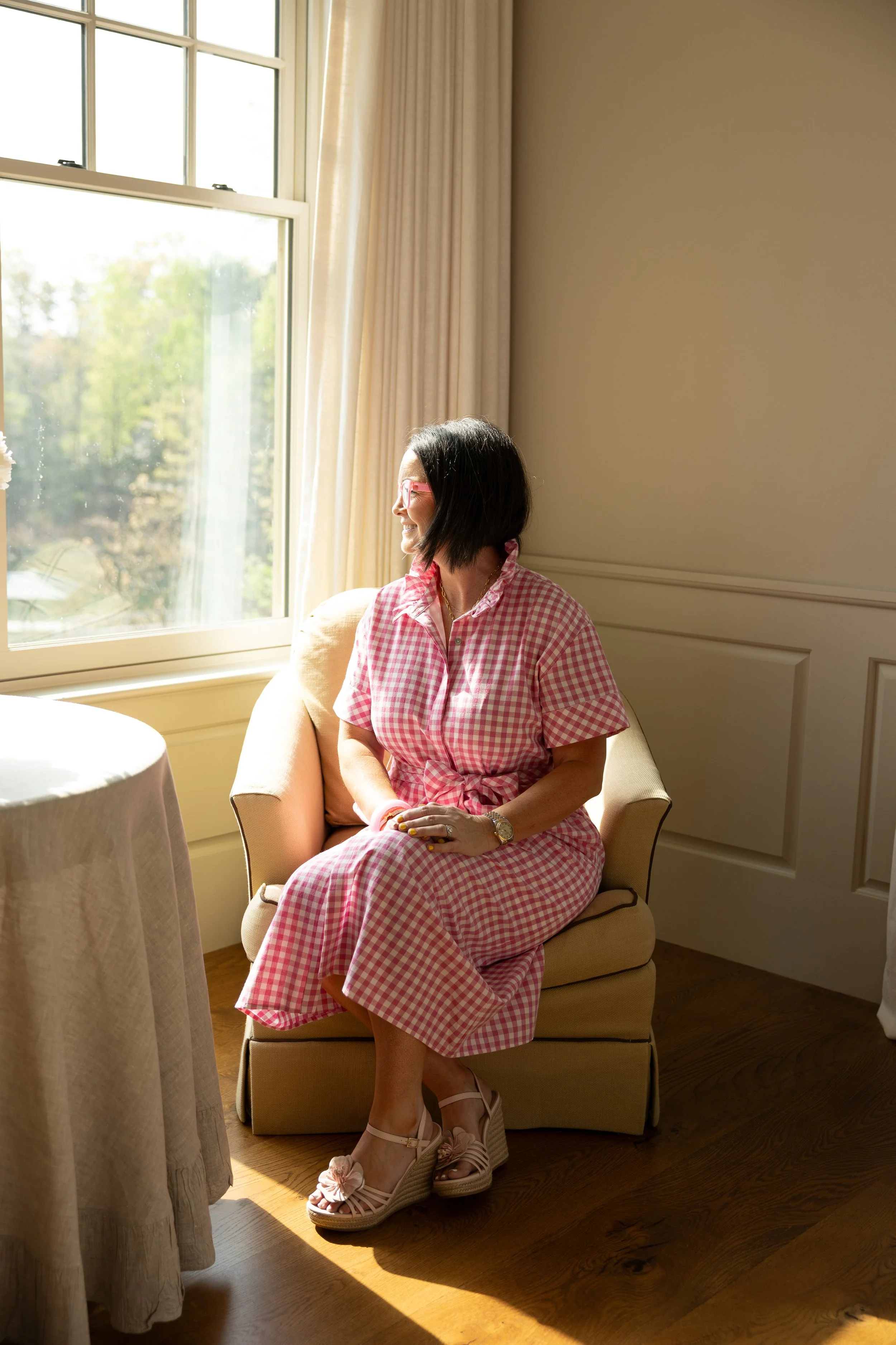 Woman wearing pink glasses and a pink and white checkered dress sitting on a beige chair by a window, smiling and looking outside, with sunlight illuminating her face and hand resting on her lap.