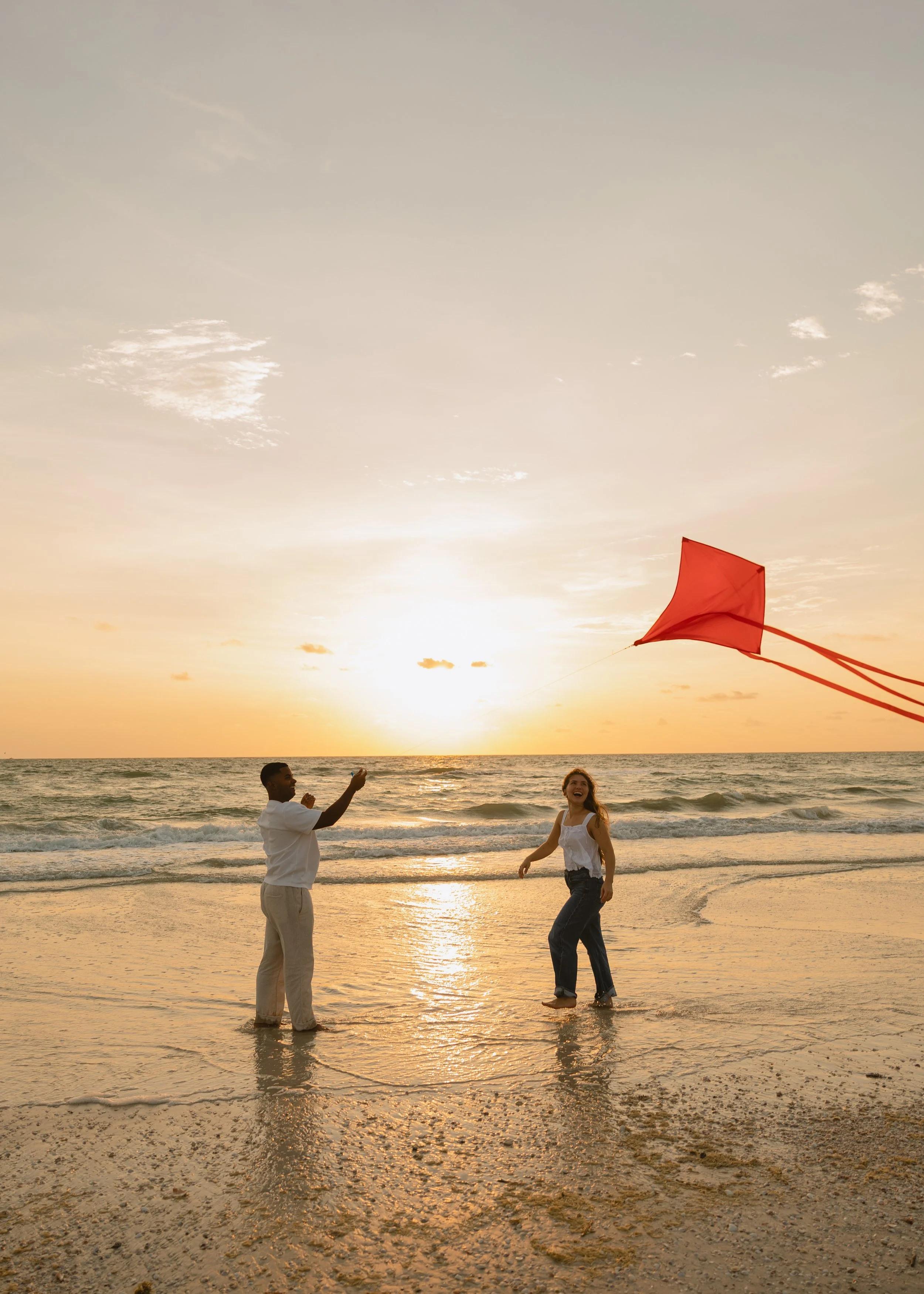 Two people are on a beach at sunset, flying a red kite while the waves gently roll onto the shore.