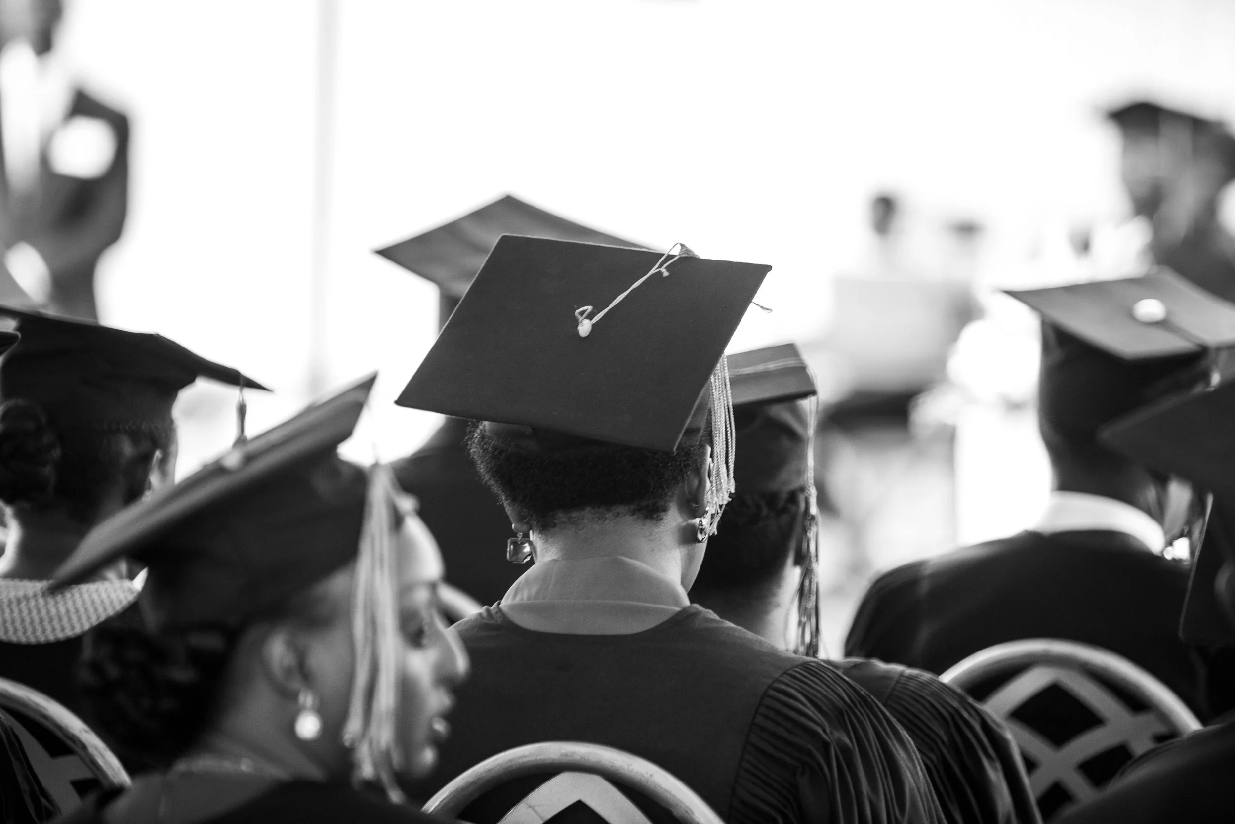 Black and white photo of graduates wearing caps and gowns attending a graduation ceremony, with many seated in rows facing forward.