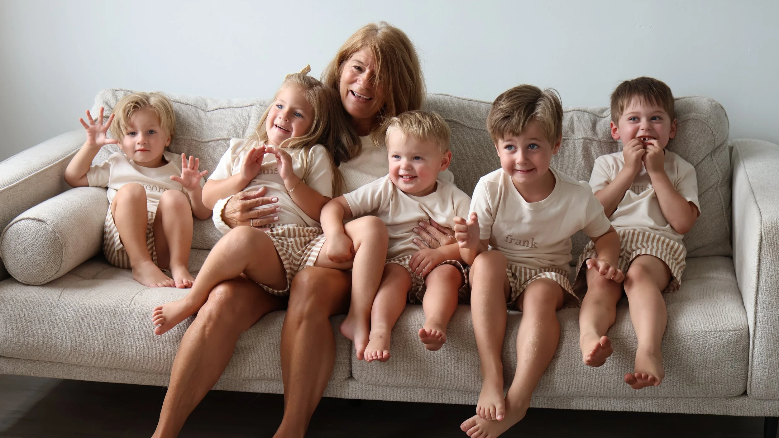 A woman with red hair sitting on a beige sofa with six young children, all wearing matching white t-shirts and striped shorts, smiling and engaging in playful interaction.