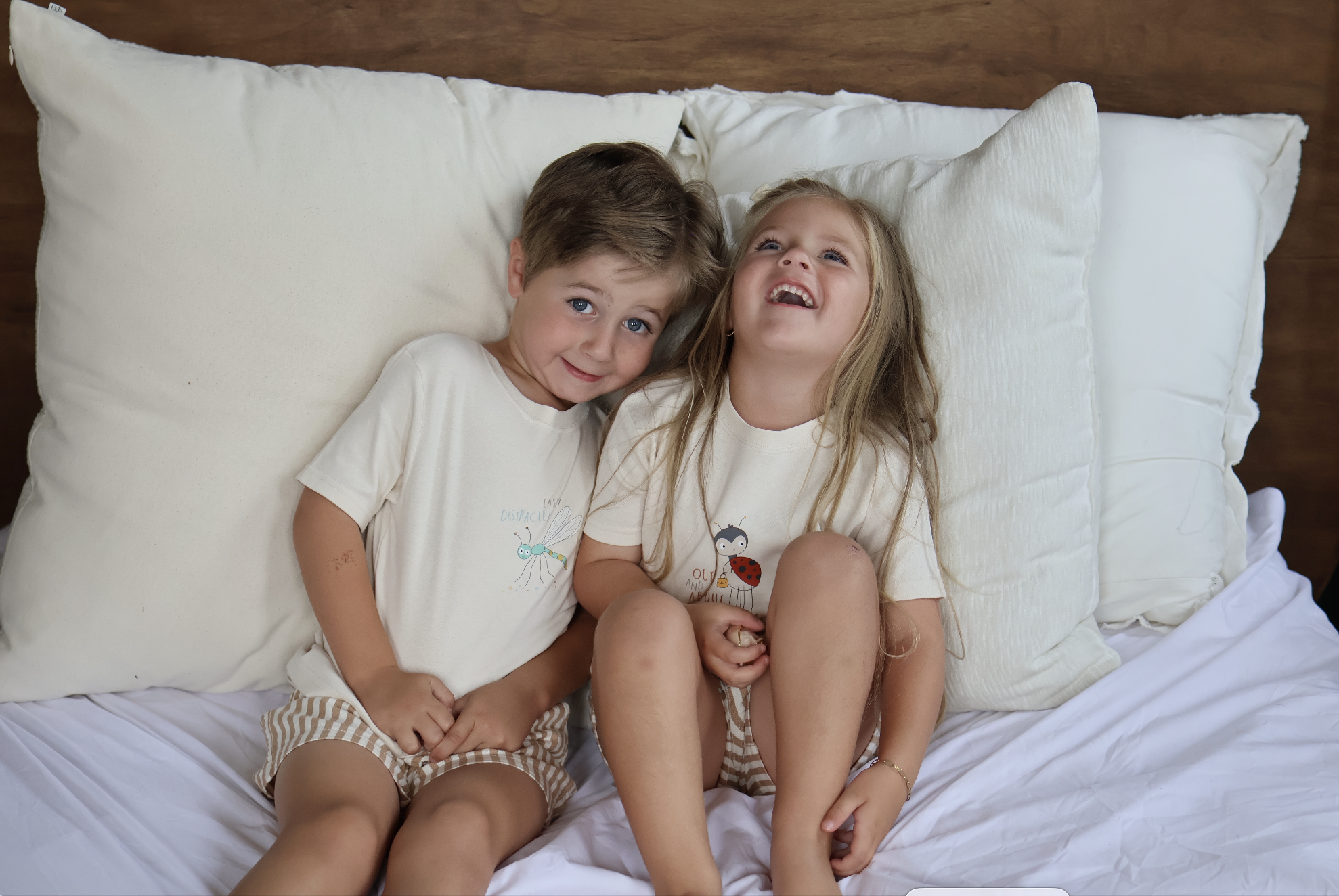 Two young children, a boy and a girl, lying in bed on white pillows, smiling and laughing. They are wearing matching pajamas with cartoon characters. The boy has short, light brown hair and blue eyes, and the girl has long, blonde hair and blue eyes.