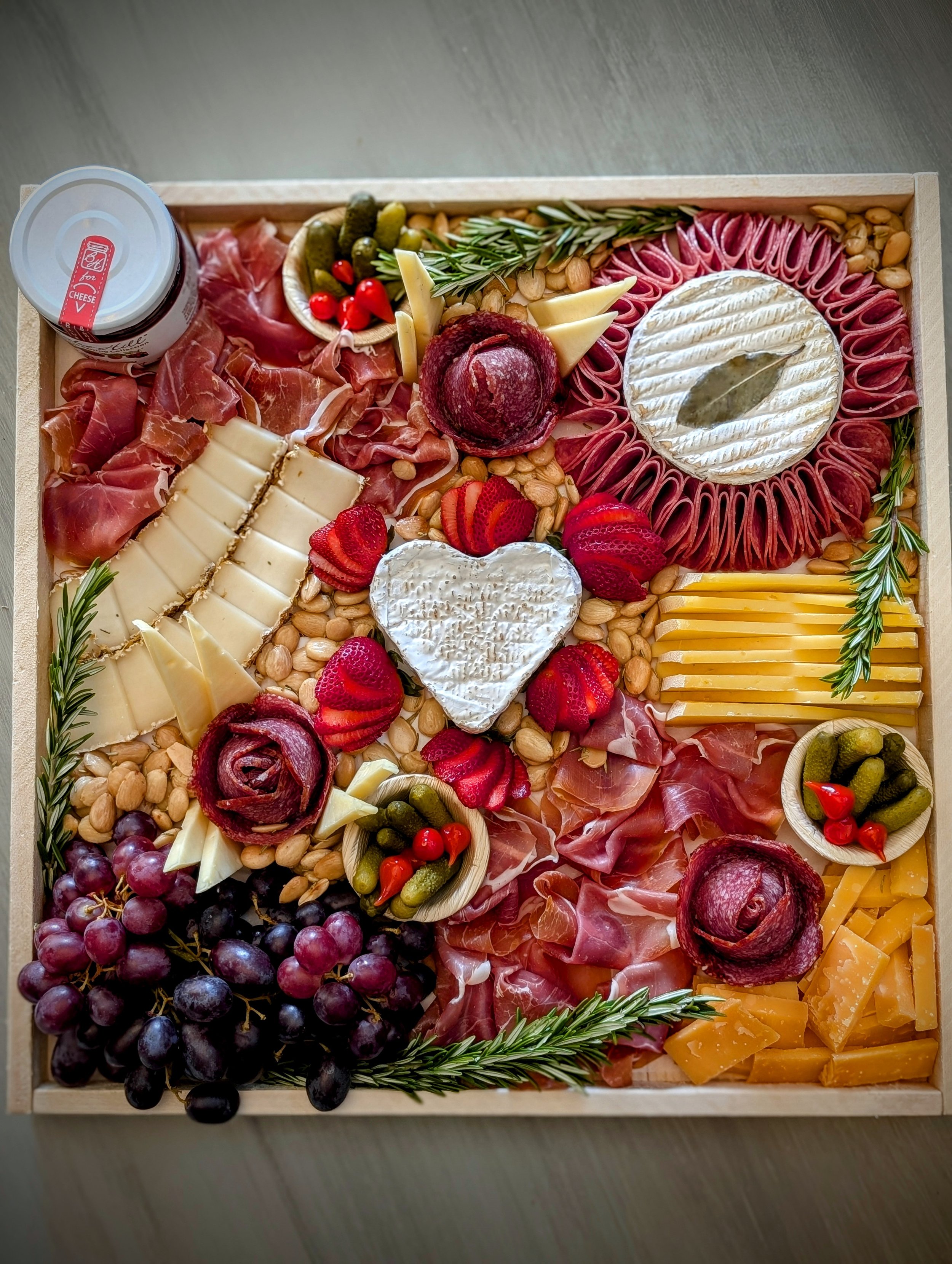 A wooden tray with an assortment of cheeses, sliced meats, grapes, strawberries, pickles, nuts, and garnished with herbs like rosemary.