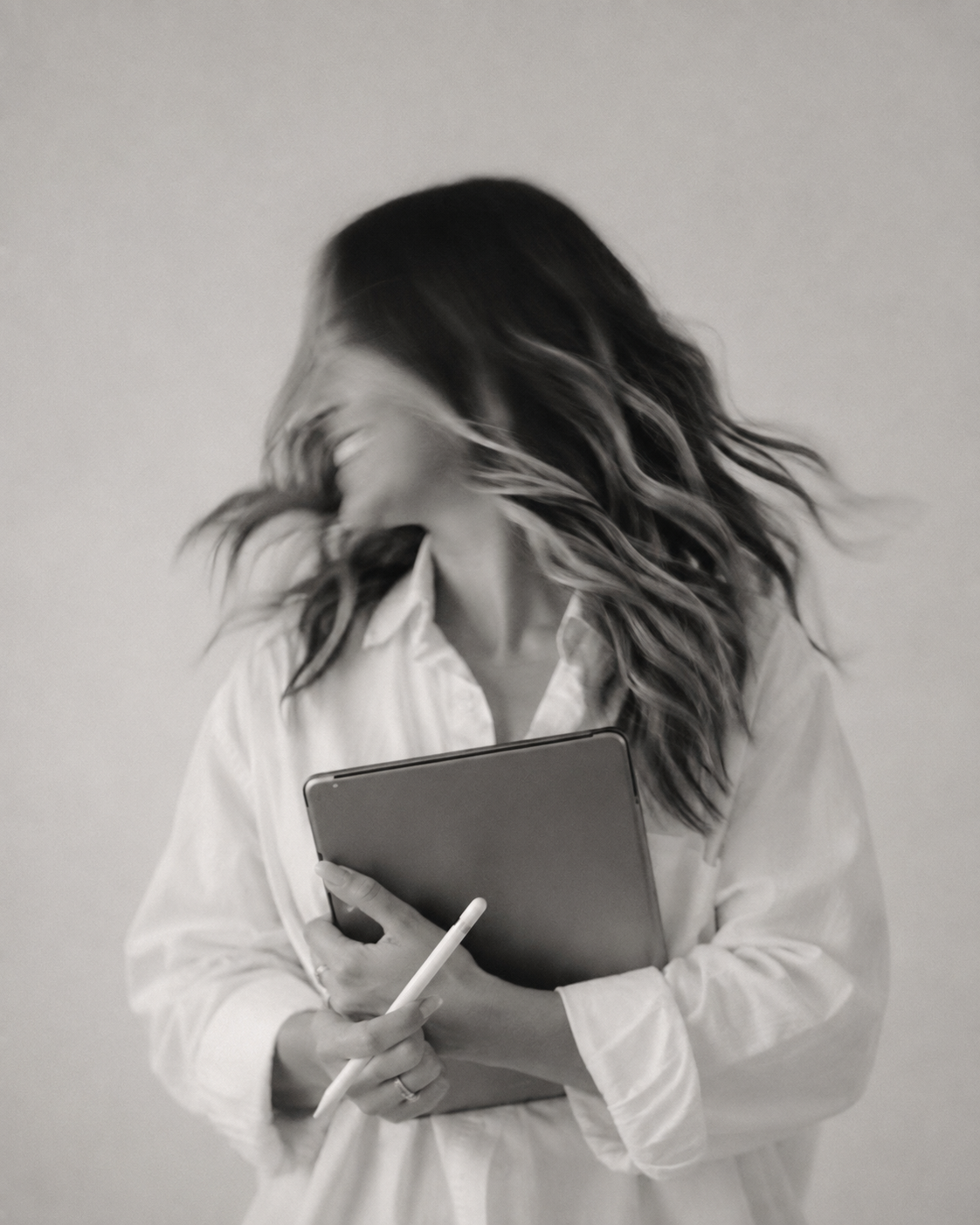 A woman with long, wavy hair moving around, wearing a white shirt, holding a tablet and a stylus, with a plain wall in the background.