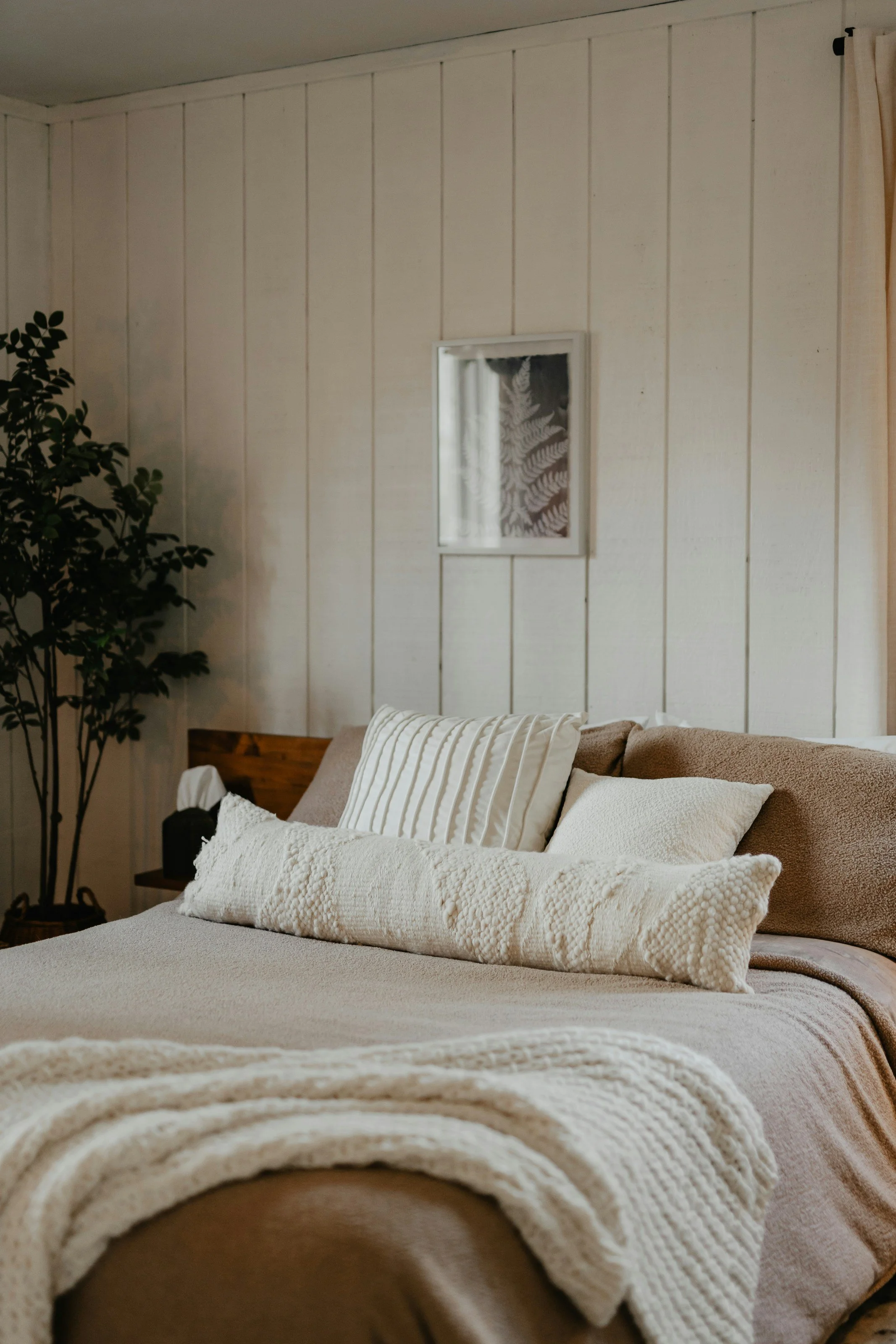 A cozy bedroom with a bed covered in beige and cream bedding, accented with multiple pillows, including a long textured cream pillow. The wall behind the bed is paneled wood, with a small framed black and white fern print. There is a houseplant to the left of the bed, and cream curtains on the right.