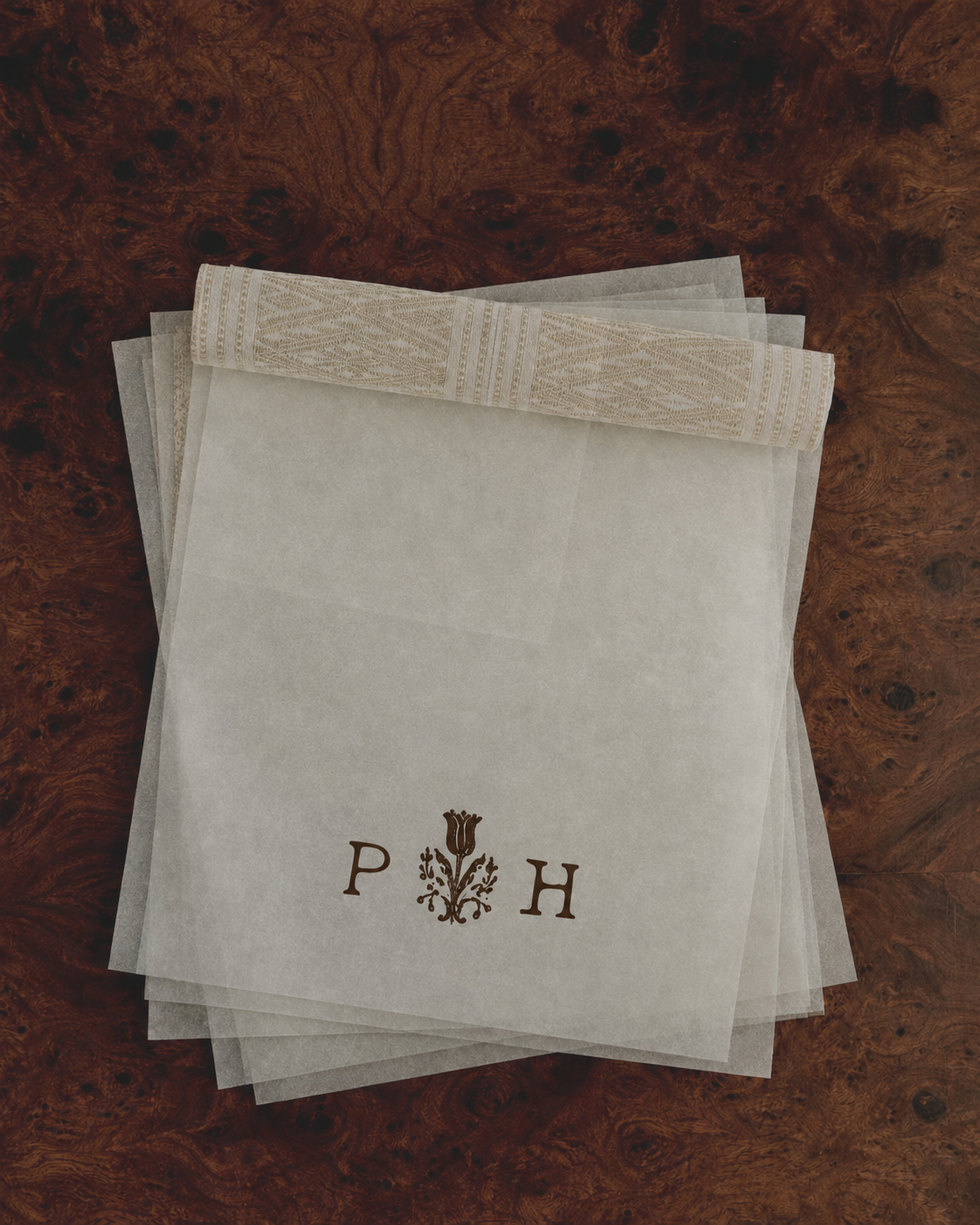 Stack of white tissue papers with an embossed floral pattern, placed on a polished wooden surface, with the initials P and H and a floral design between them on the top tissue paper.
