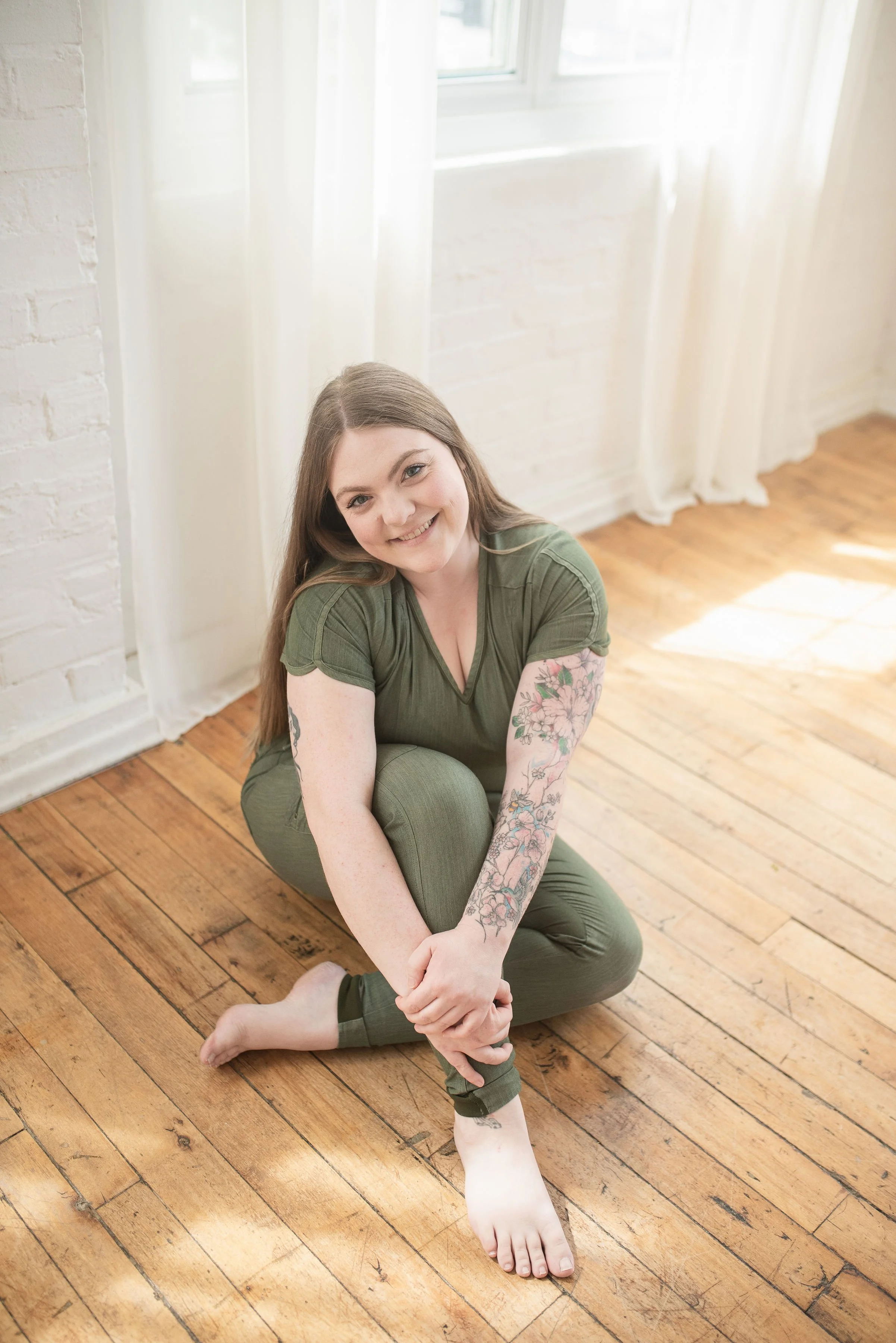 Jessi is sitting barefoot on a hardwood floor, smiling at the camera and wearing green medical scrubs