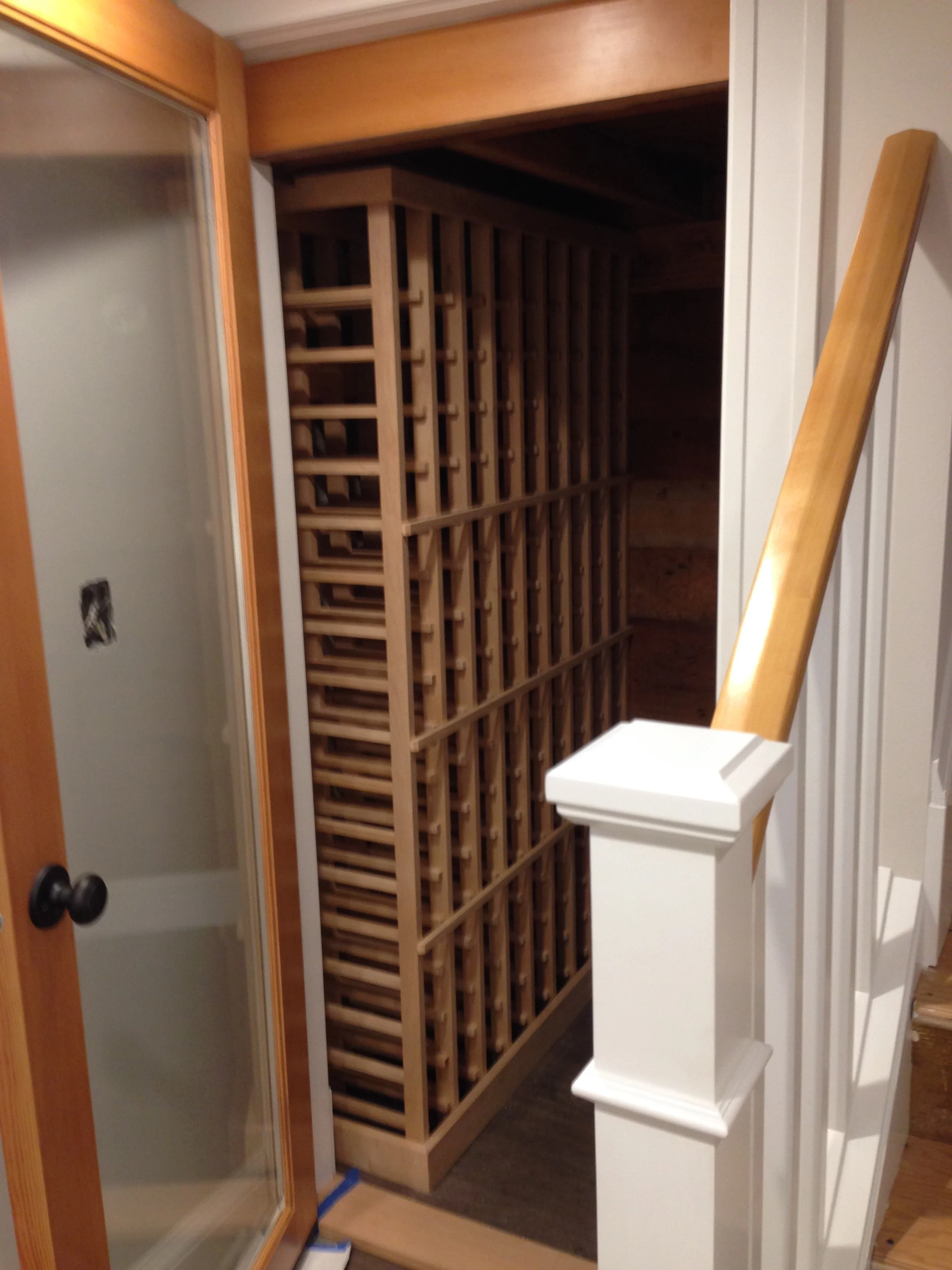 A wooden wine rack inside a partially built or renovated room. Wine cellar with custom racks and wine cooler with humidifier. glass door installed. Painted walls and stair trim painted.