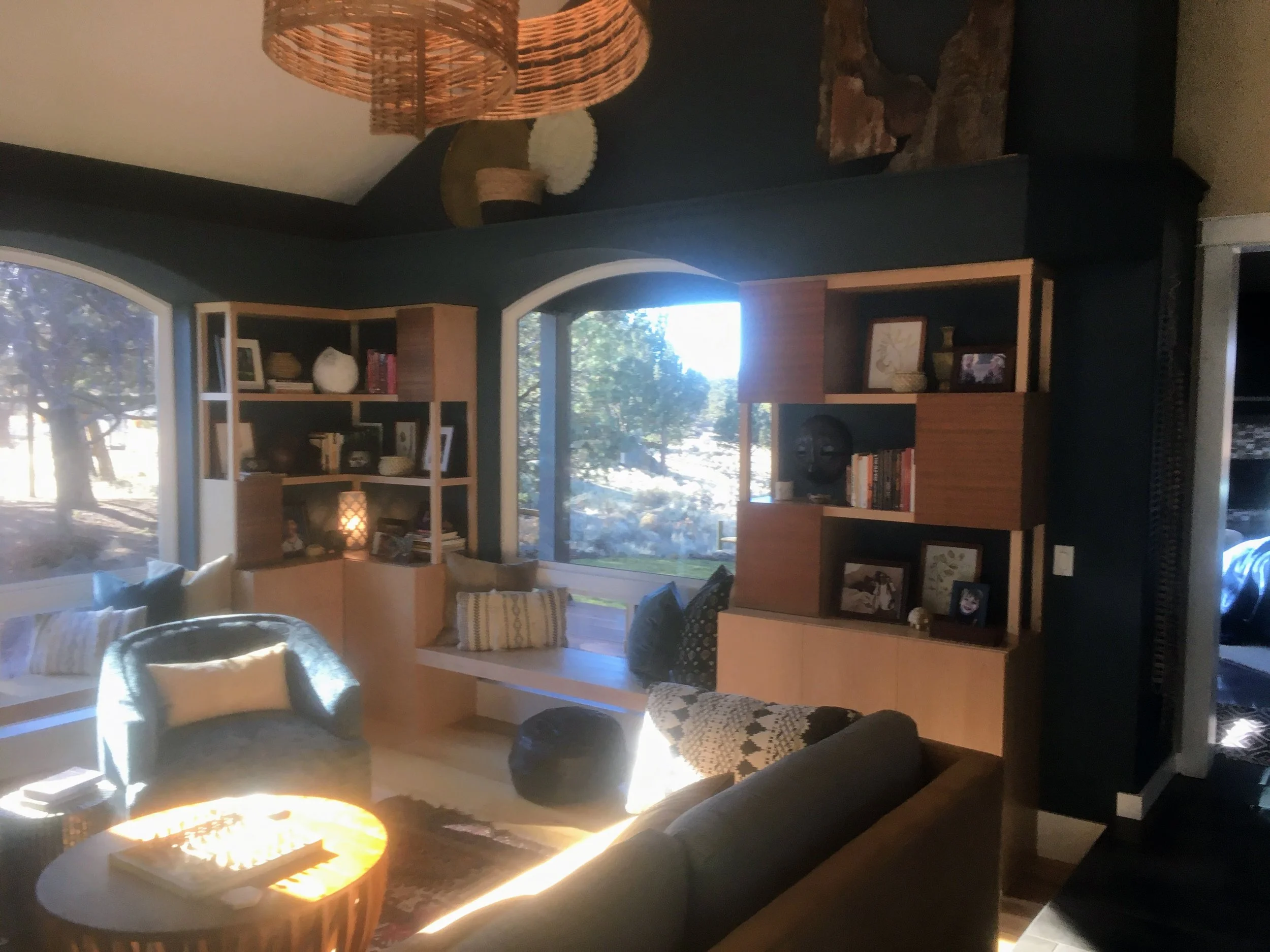 Living room with dark walls, large windows installed, modern bookcase with window seat built between and decorative items and books, cozy seating area with armchairs and pillows, fireplace with stones, and a round coffee table with a lit candle.