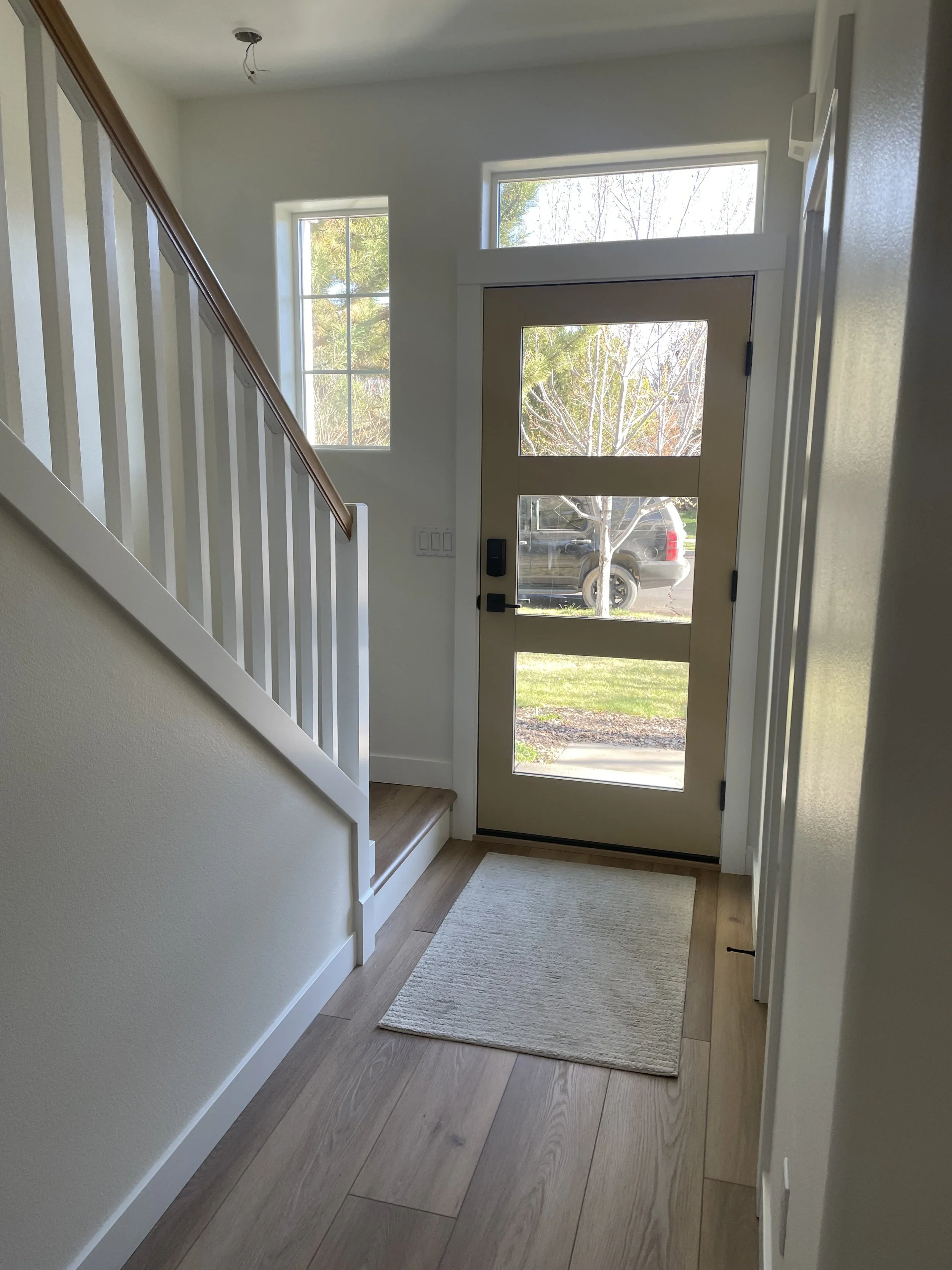 A small entryway with light-colored walls, a wooden staircase railing, a beige front door with large glass panels, a view of a neighborhood outside, and a white rug on the wooden floor. New LVP flooring installed. Modern front door installed.