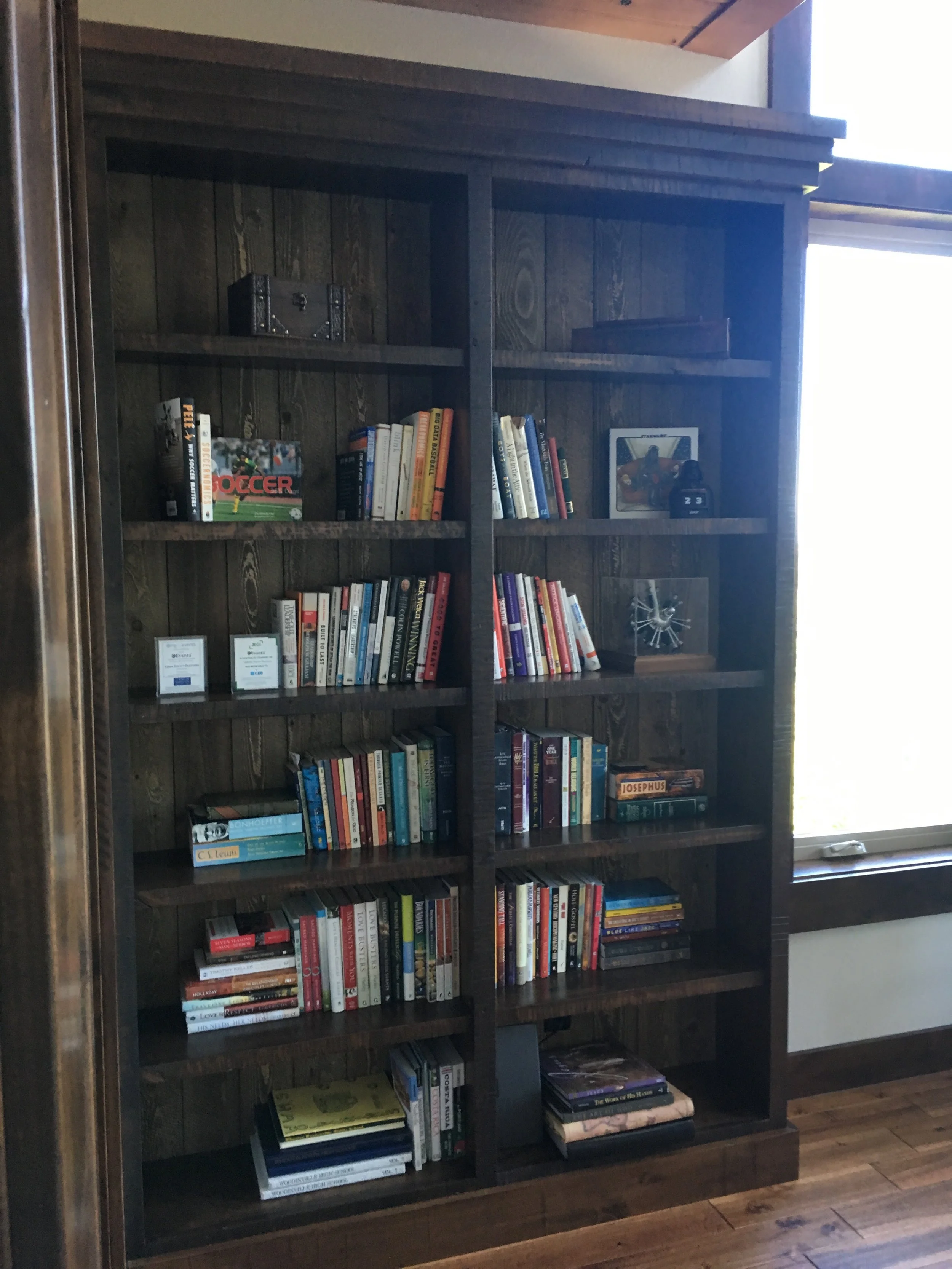 A dark wooden bookshelf filled with various books and some decorative items, located next to a window with natural light streaming in.