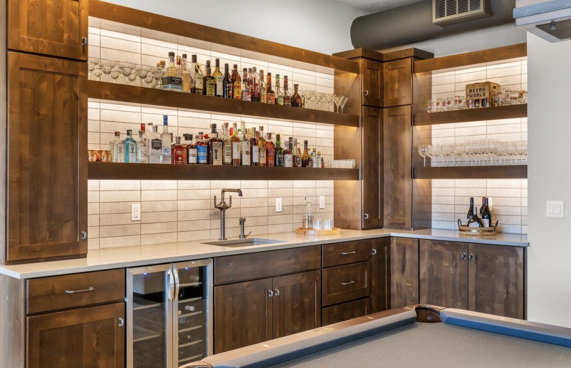 A home bar with wooden cabinets, open shelves displaying various liquor bottles and glassware, a wine refrigerator, a bar sink, and a countertop with a tray of glasses and a tray with bottles, against a tiled backsplash.