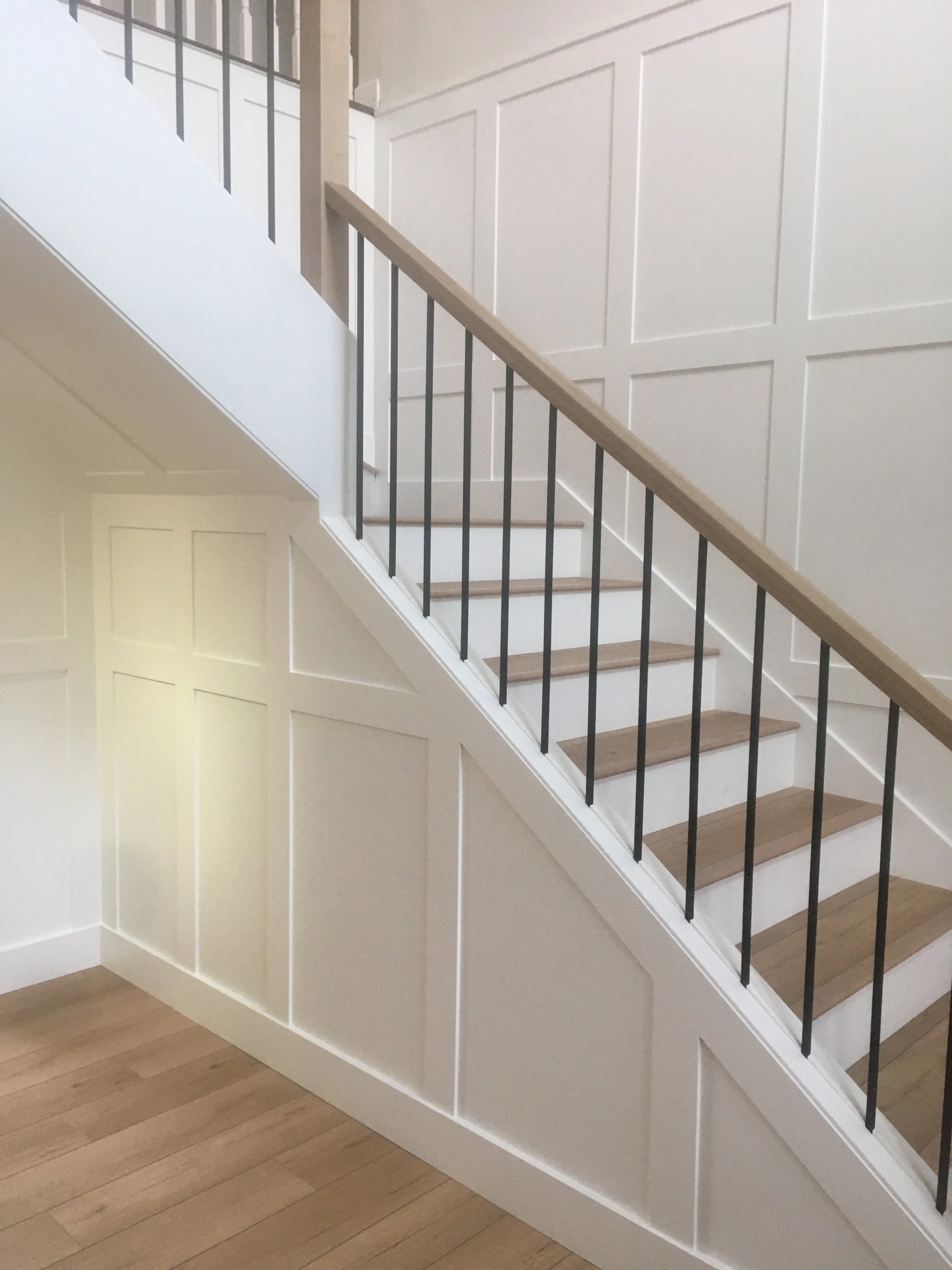 Interior view of a staircase with wooden steps, white paneling on the wall, and a black metal railing.