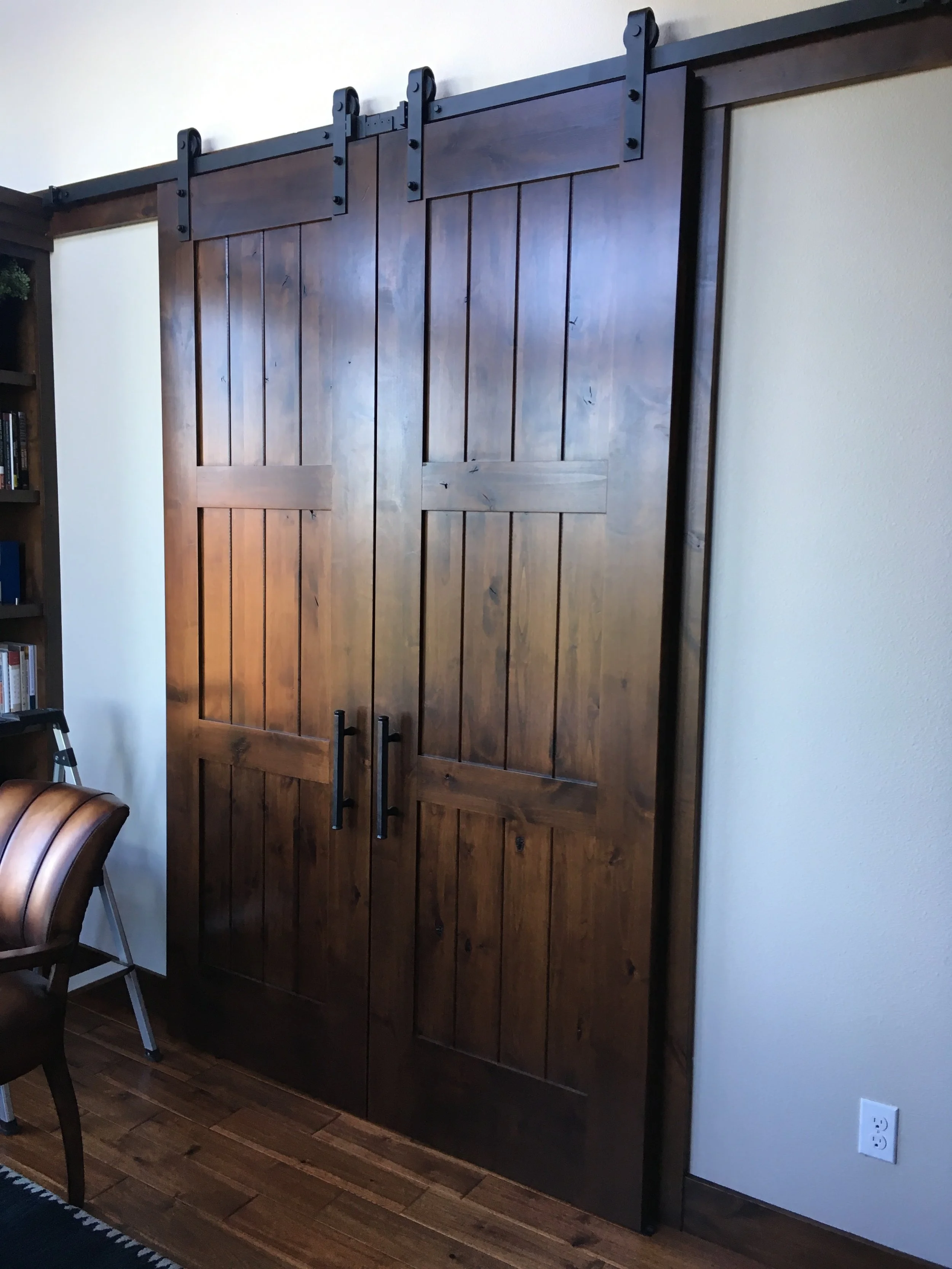 Wooden sliding barn door with black metal hardware, mounted on a white wall in a room with wooden flooring.