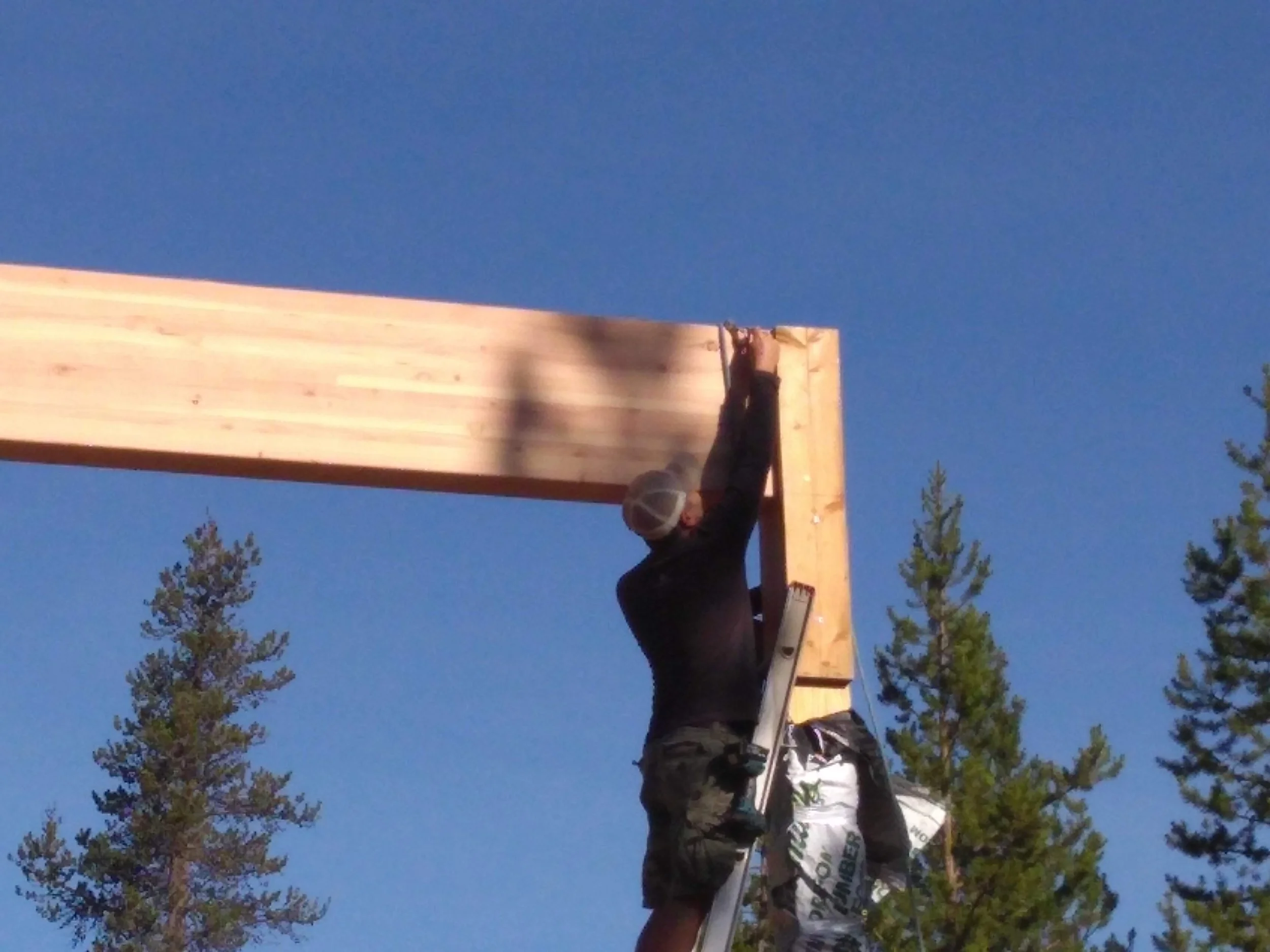 A person wearing a hat and camouflage clothing is working on a wooden structure outdoors against a clear blue sky, with trees in the background.