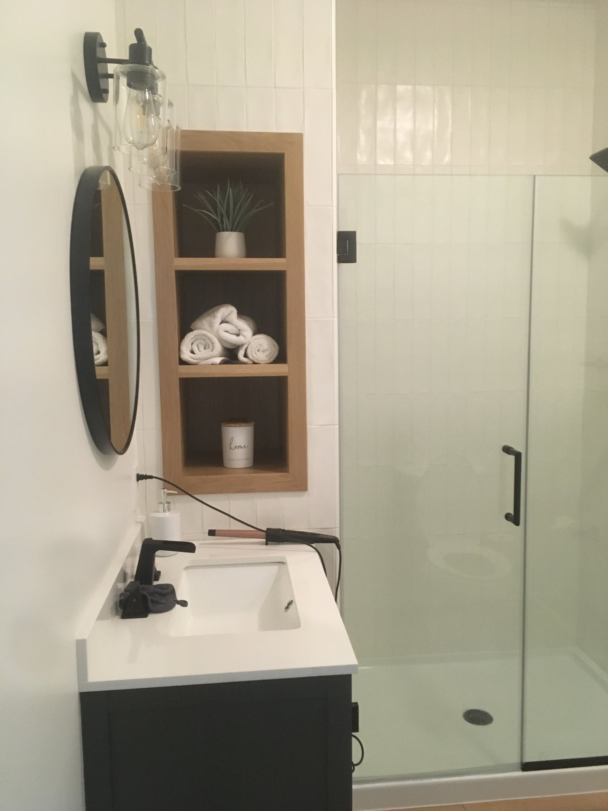 Bathroom with white sink, black cabinet, oval mirror, wooden shelf with potted plant and rolled towels, glass shower enclosure, and a showerhead.