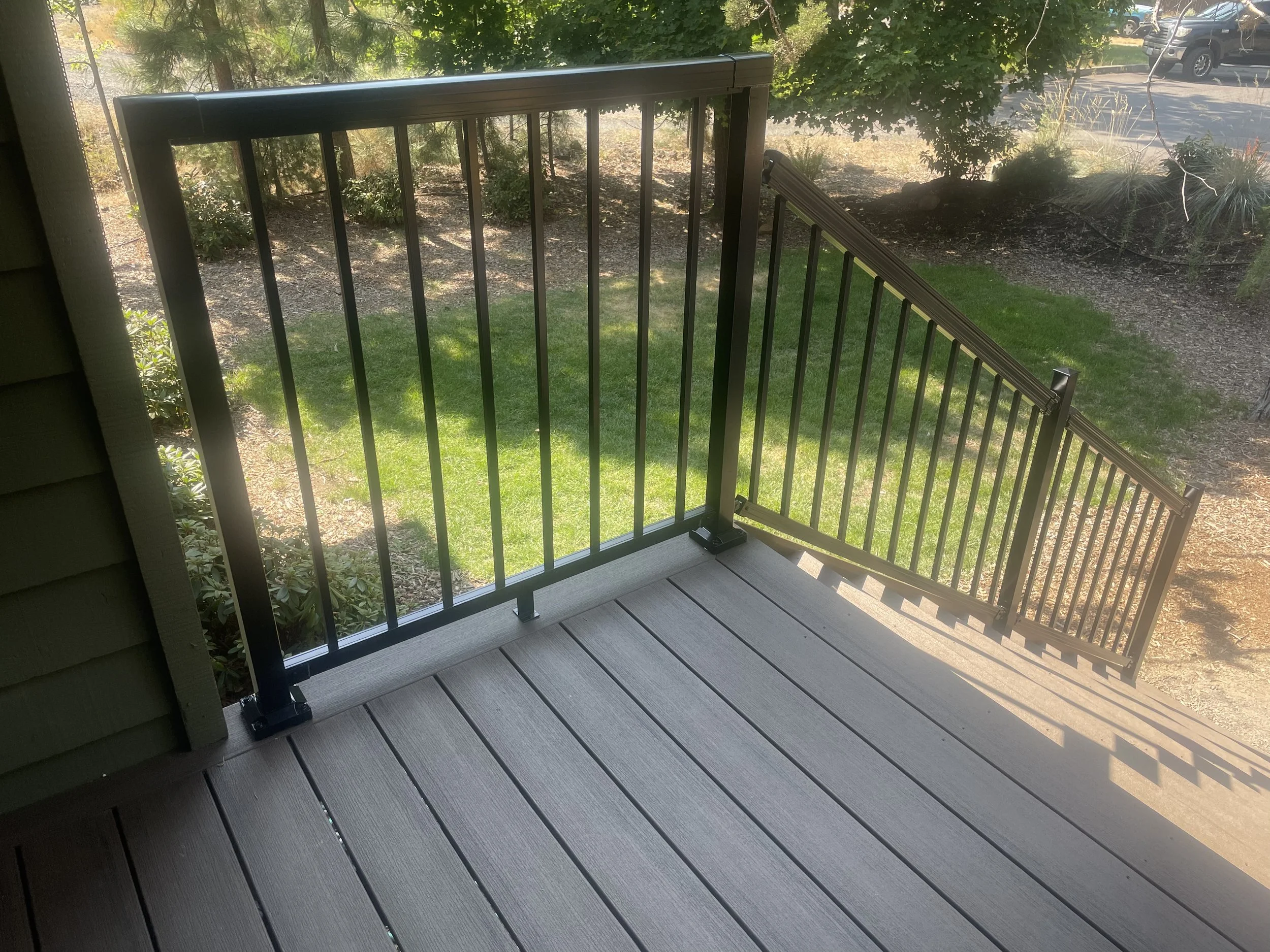 View of a backyard from a wooden porch with black metal railings, grass, bushes, trees. composite decking trex and Timbertech