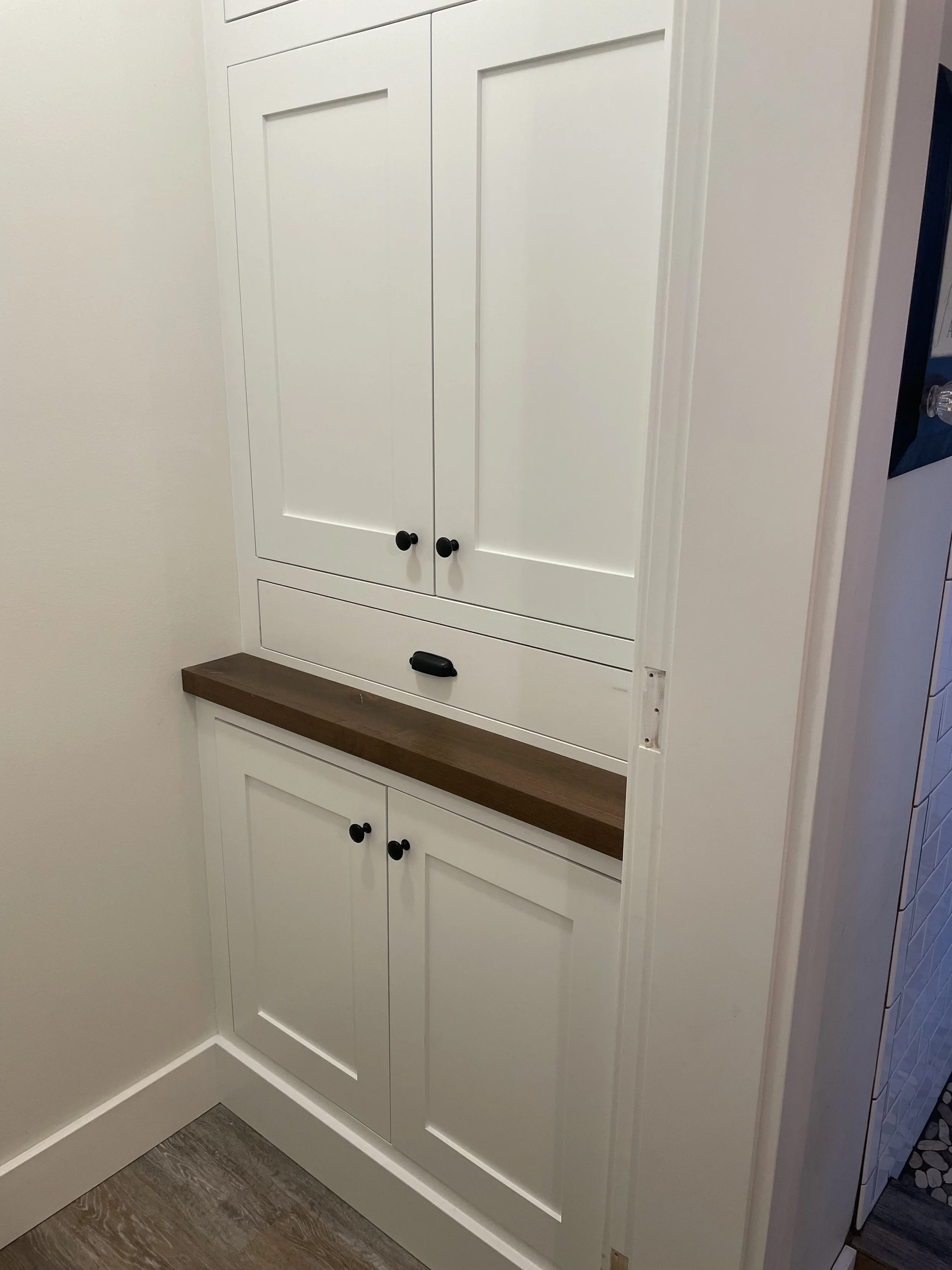 White kitchen cabinet with black knobs, part of a kitchen with tiled floor and a wooden shelf below the cabinet.