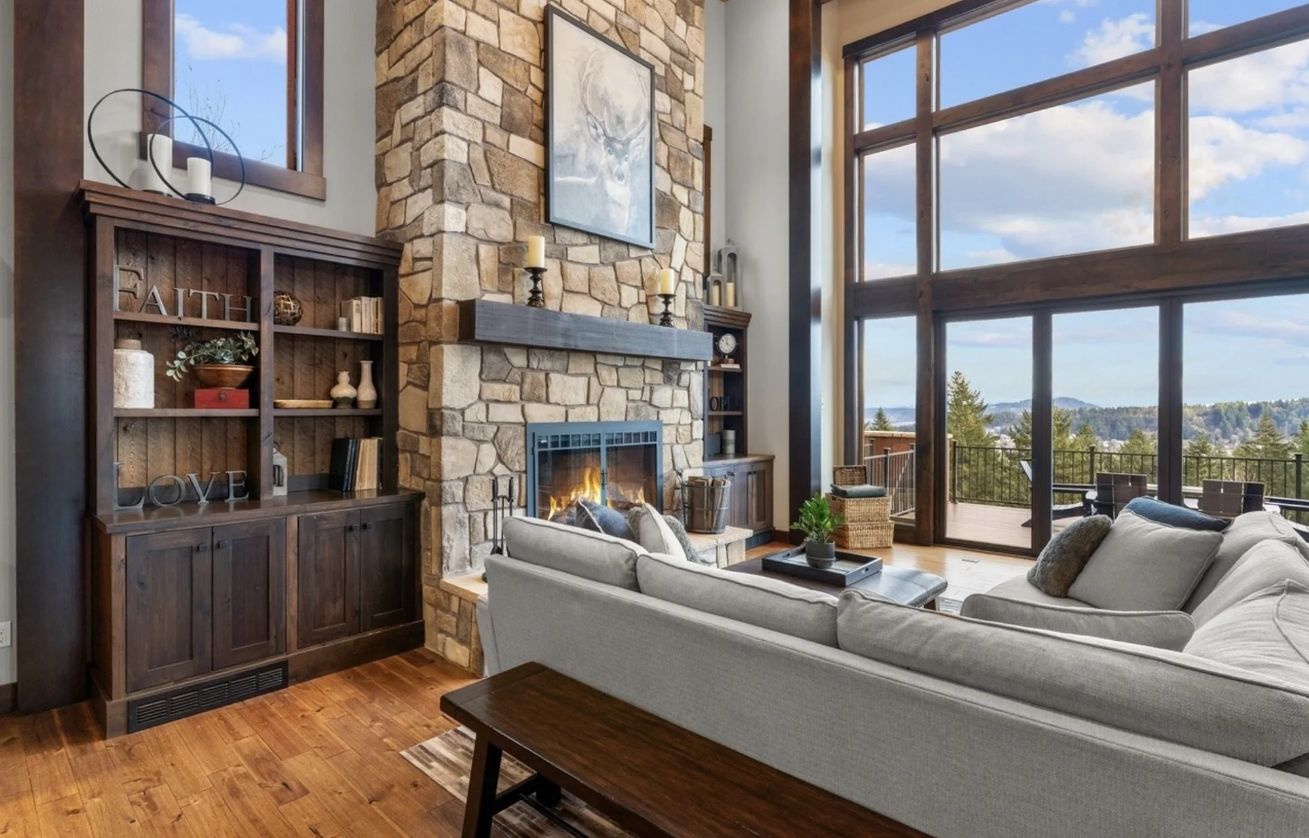 Living room with a stone fireplace, wooden shelves with decorative items, a large window with a view of trees and mountains, and a cozy seating area with a sofa and armchair.