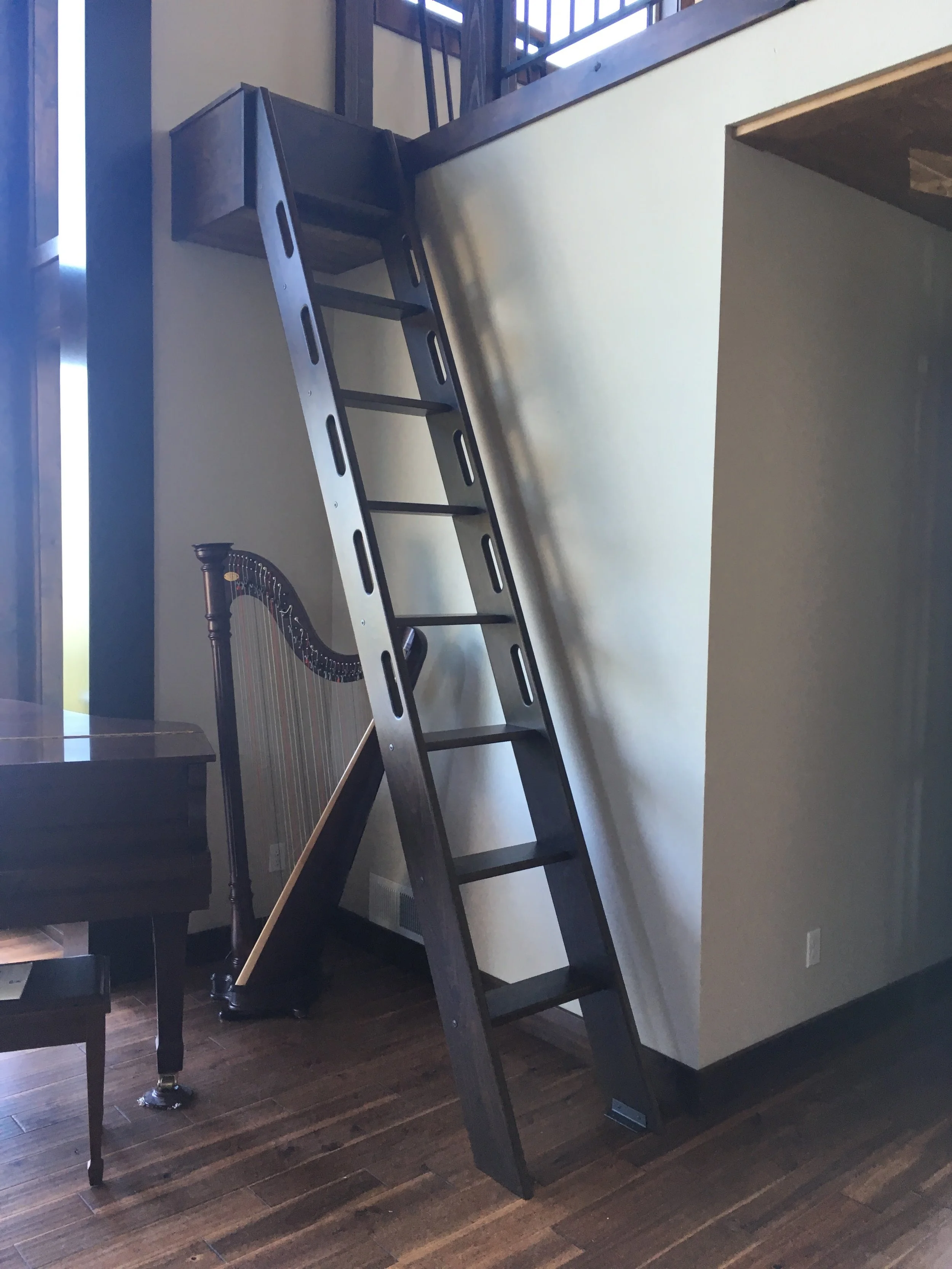 A black wooden ladder leaning against a white wall in a room with dark hardwood flooring, with a harp visible in the background.