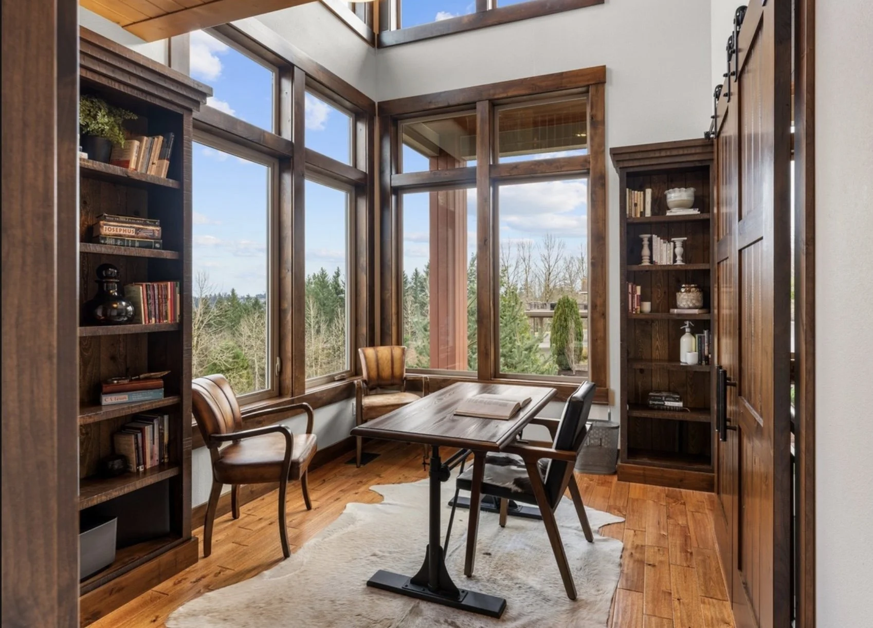A cozy corner home office with large windows, wooden shelves filled with books, a wooden desk, and two chairs. There is a carpet on the wooden floor and a cowhide rug, with a view of trees and sky outside.