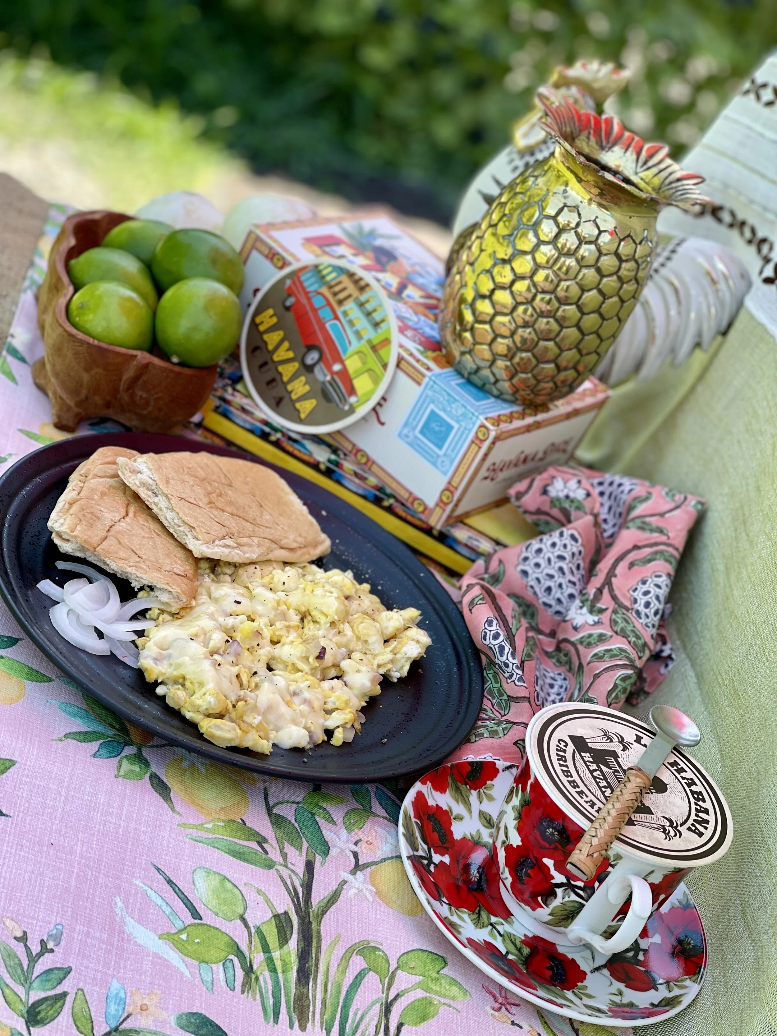 A table set with a floral tablecloth, holding a plate of macaroni salad with onions and a slice of bread, a bowl of green grapes, a decorative yellow pineapple-shaped container, a box of crackers, a container of cherry butter, and a napkin with a flo