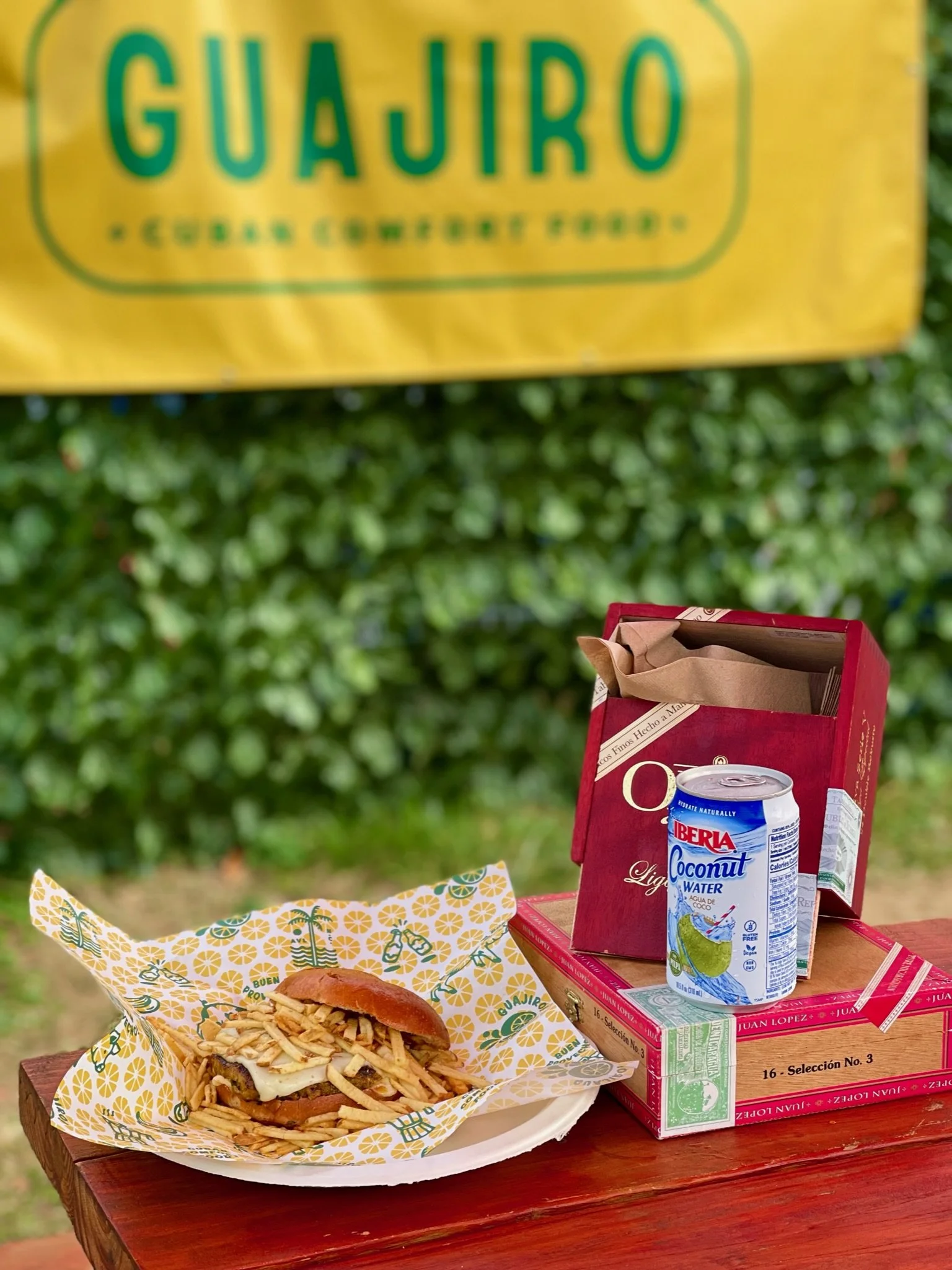 A tray of food with a burger and fries, a can of Iberia coconut water, and a red box of Juan Lopez cigars on a wooden table outdoors, with a yellow Guajiro sign hanging above and a background of green bushes.