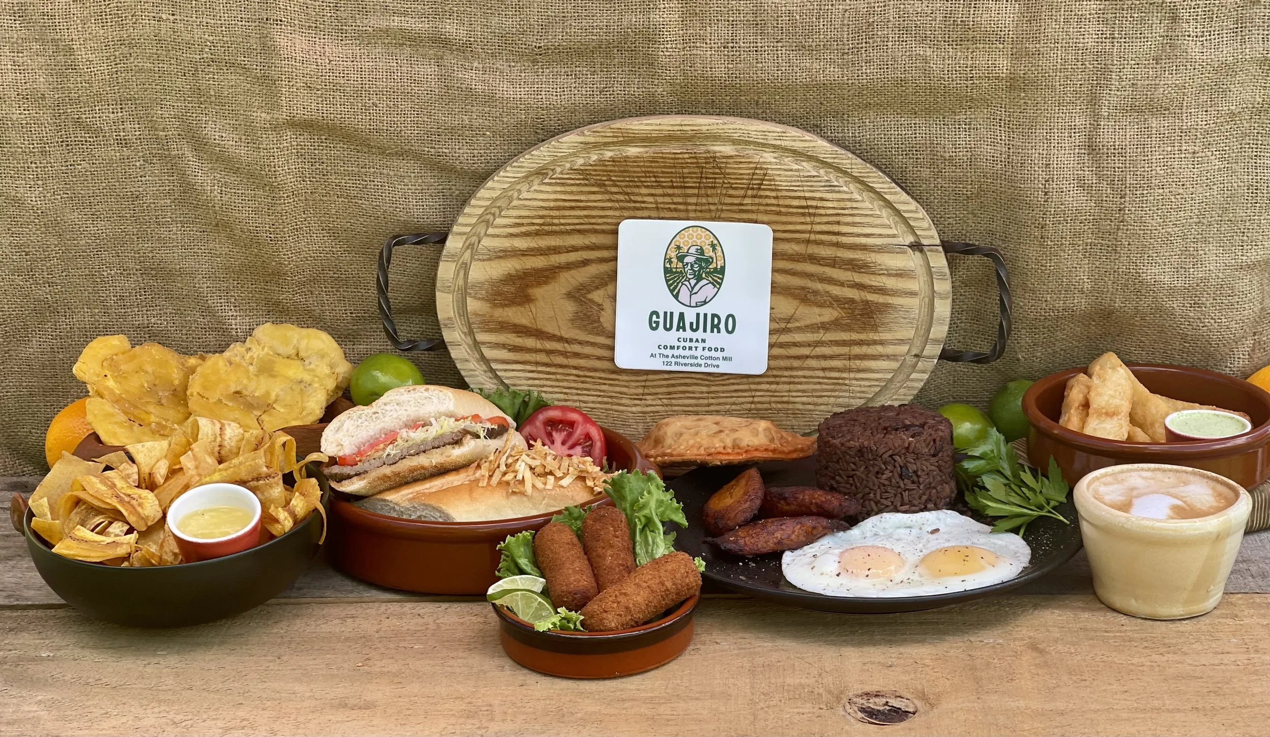 Assorted Latin American dishes including rice, fried eggs, plantains, tacos, fried plantains, salsa, and fried appetizers, displayed on a rustic wooden table with a burlap backdrop.